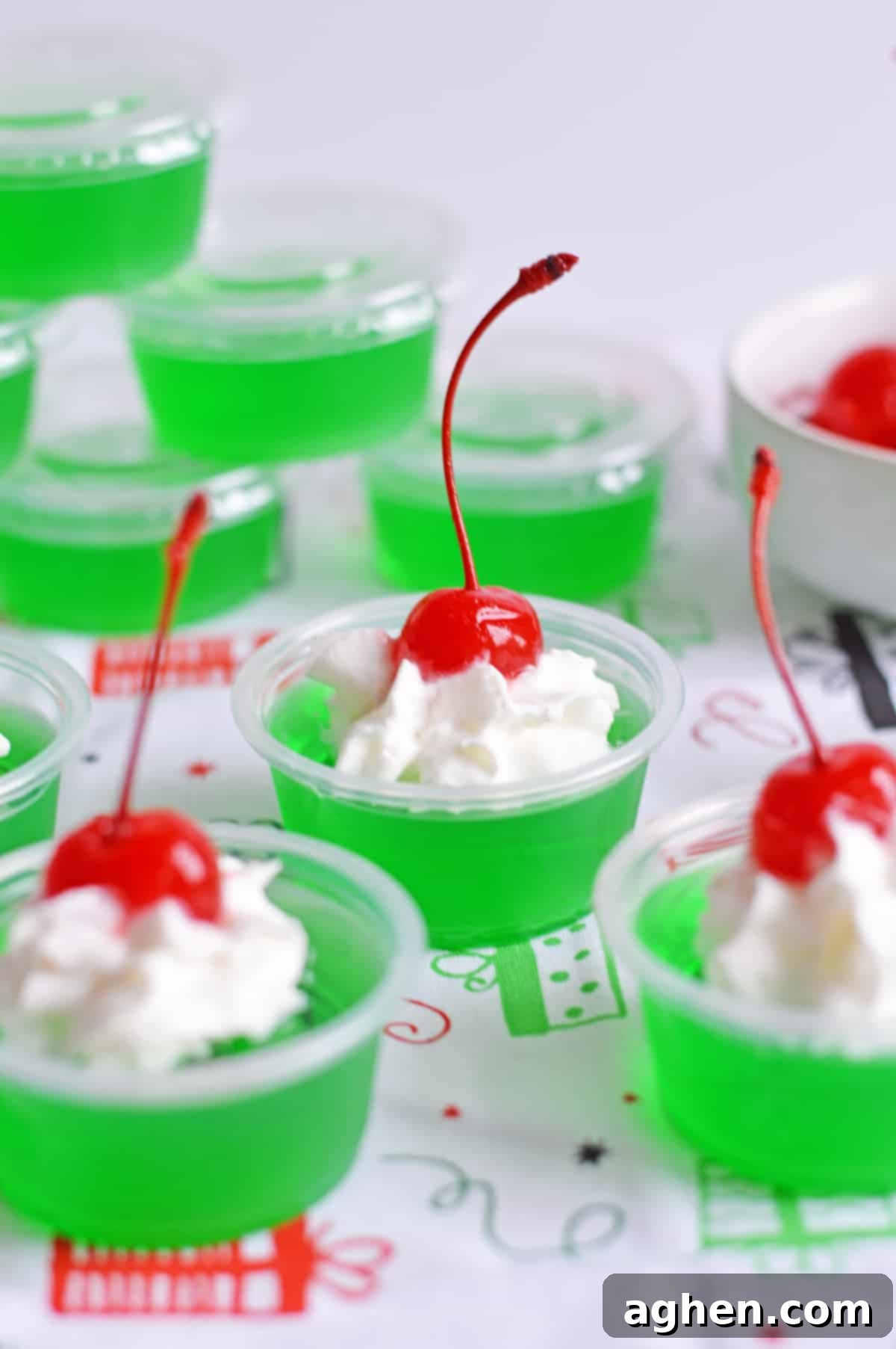 Grinch Jello Shots in small plastic shot cups made with lime jello, tequila, whipped cream, and a cherry on top