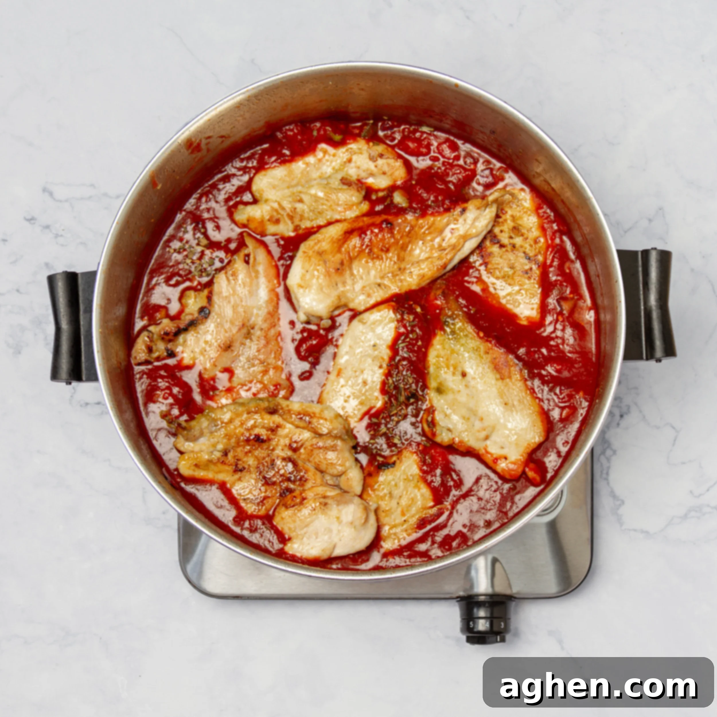 A close-up view of sautéed vegetables and perfectly seared chicken breasts simmering together in a rich, vibrant tomato sauce within a large pan. The sauce is bubbling gently, and the ingredients are melding, creating a deep, complex flavor for the Chicken Creole.