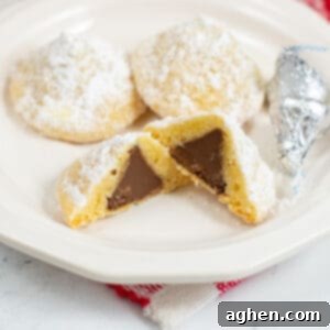 Hidden kiss cookie cut in half to show hershey kiss in center. Two whole cookies and 2 wrapped hershey kisses are next to the cookie on the plate.