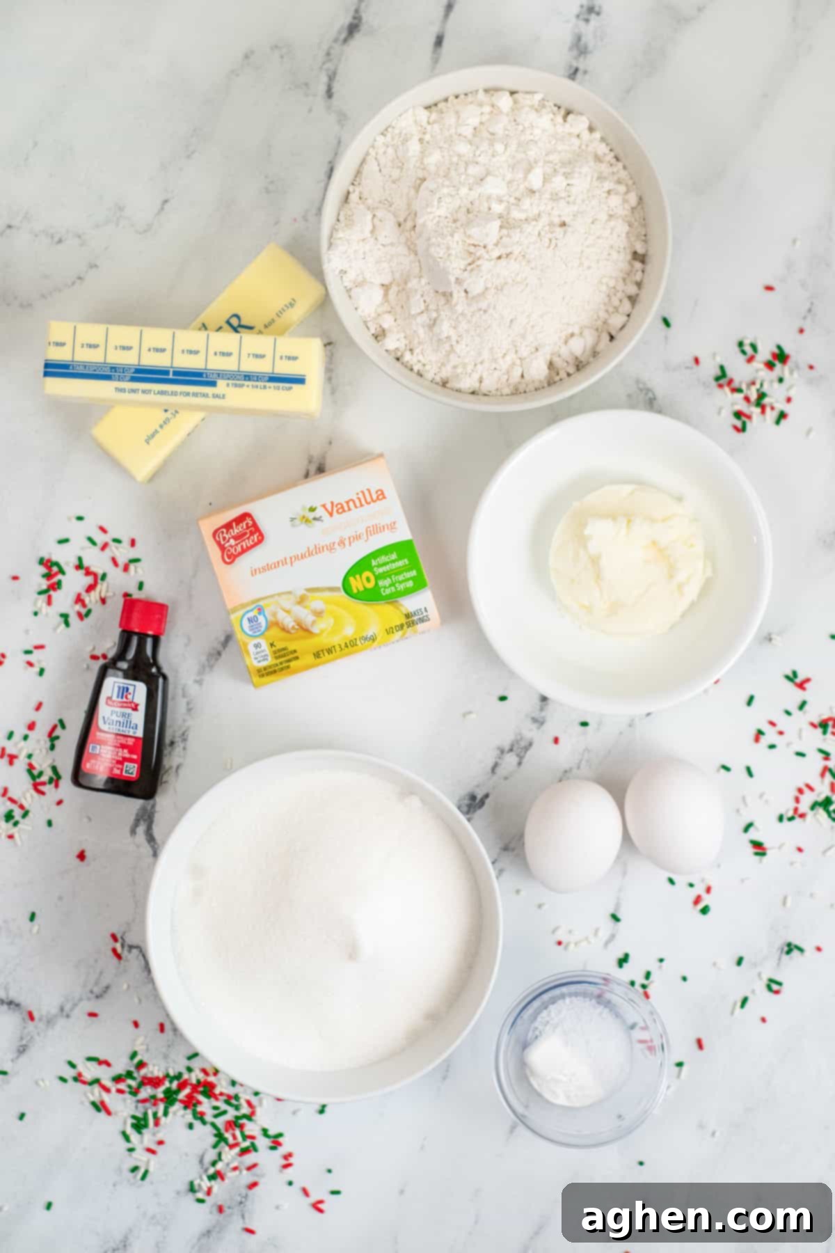 Two sticks of butter, bowl of flour, bowl of shortening, 2 eggs, box of vanilla pudding mix, bowl of granulated sugar, baking powder, bottle of vanilla, and red, white, and green sprinkles scatted all around