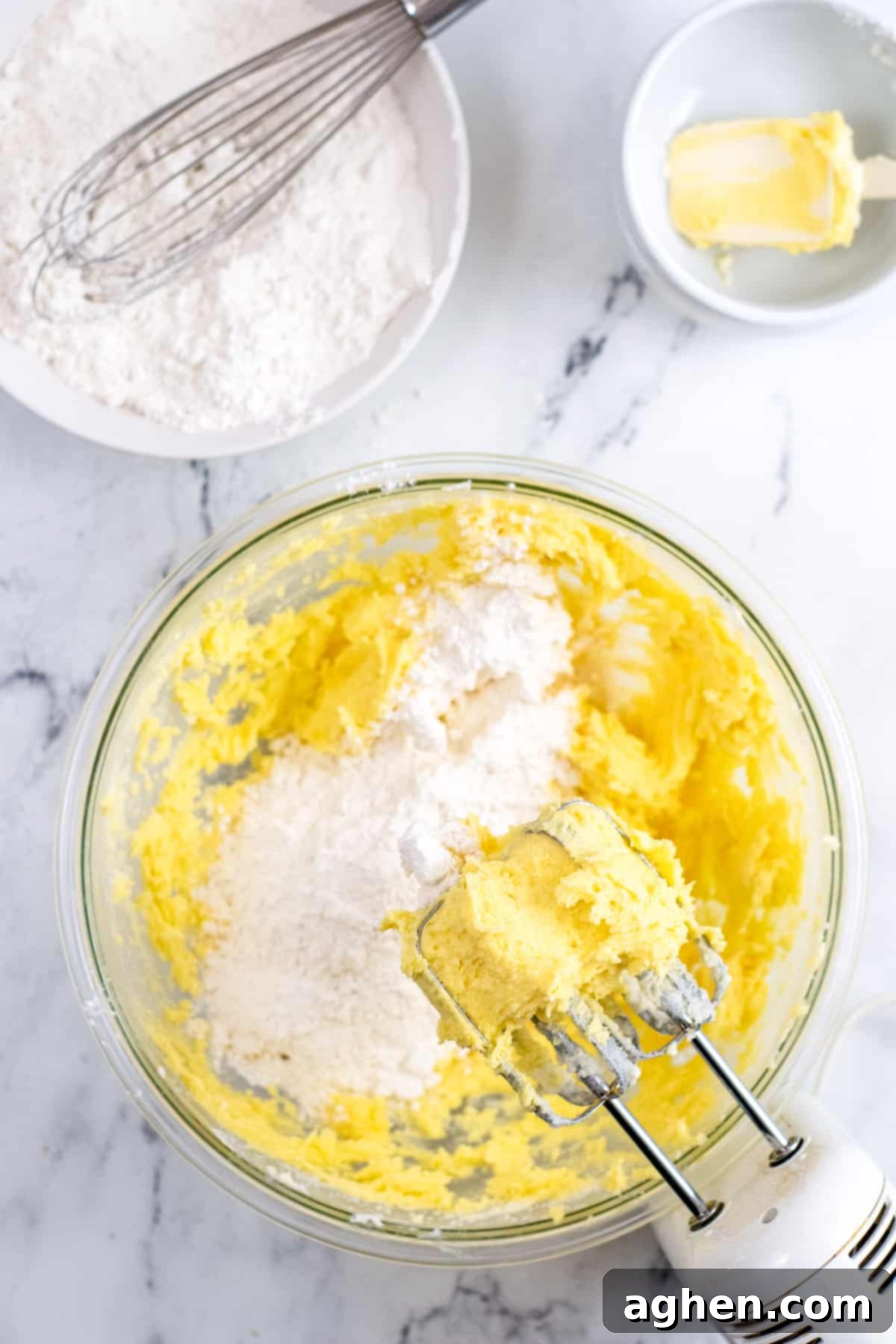 Handheld electric mixer on the side of a large mixing bowl with butter mixture and dry ingredients being gradually added.