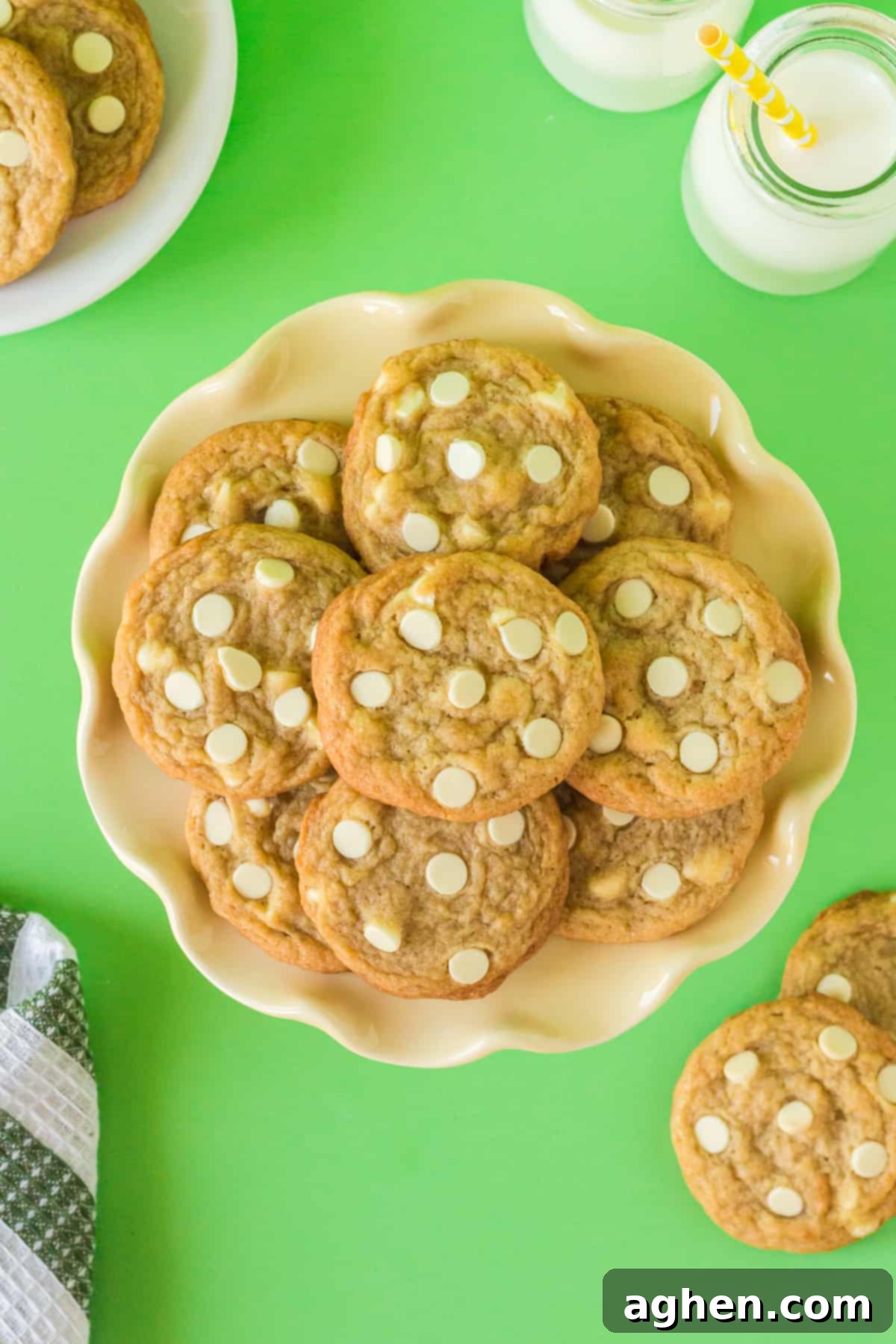 Banana pudding cookies with white chocolate chips on plate with glasses of milk and additional cookies in background.