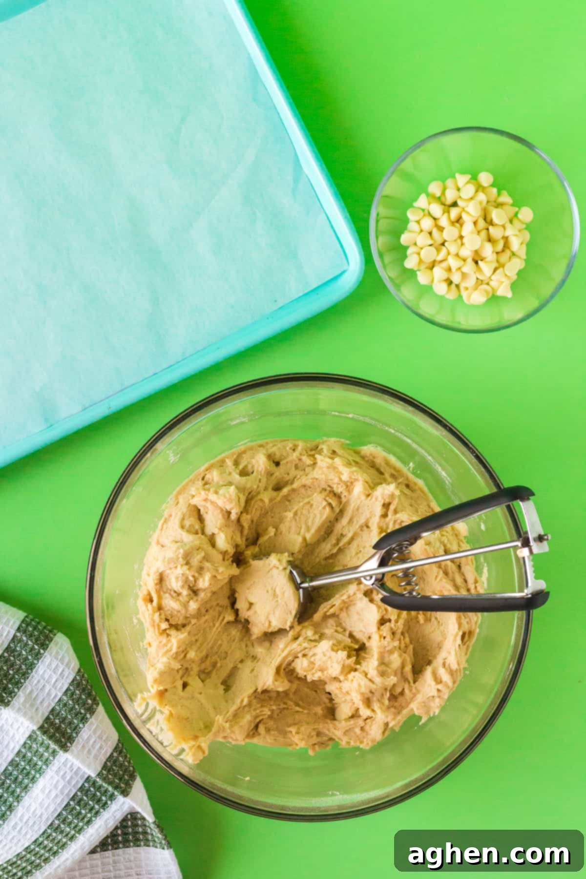 Cookie dough being scooped with a cookie scoop. Lined baking sheet and white chocolate chips are next to the bowl.