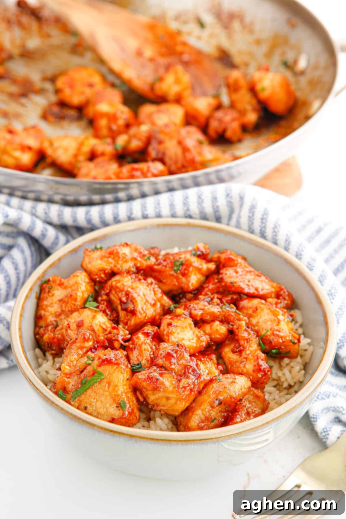 Sticky garlic chicken bites with a sweet and spicy sauce served over rice in a bowl. A skillet with additional chicken pieces and a wooded spoon is in background.
