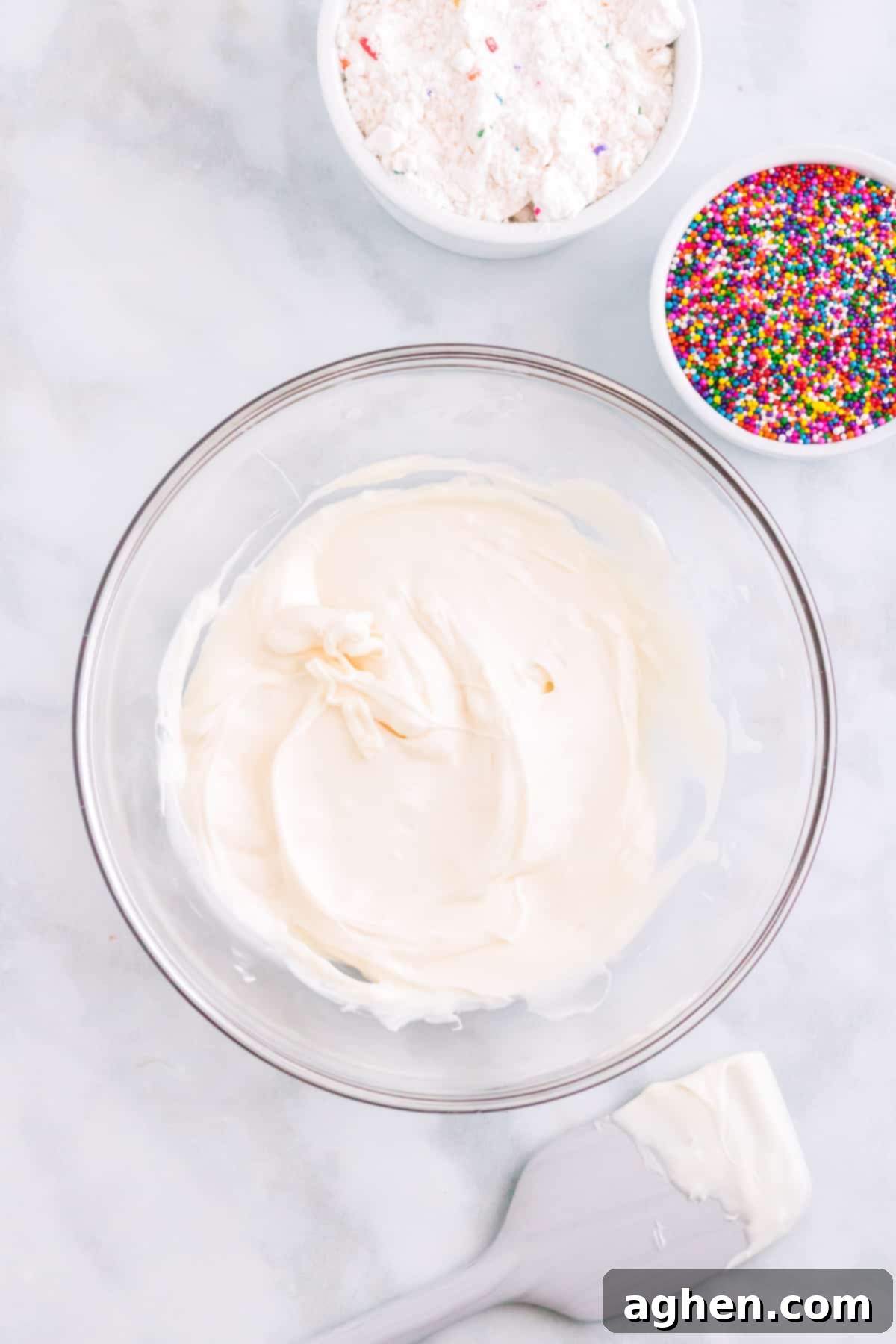 Melted white candy melts in glass bowl with rainbow sprinkles, white chocolate covered spatula, and funfetti cake mix next to it