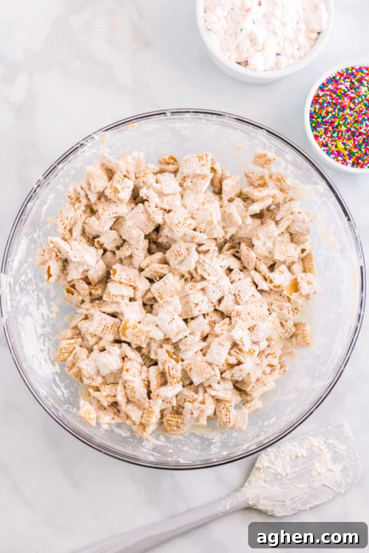 White candy melt coated chex cereal in large glass bowl with rainbow sprinkles, spatula, and funfetti cake mix beside it