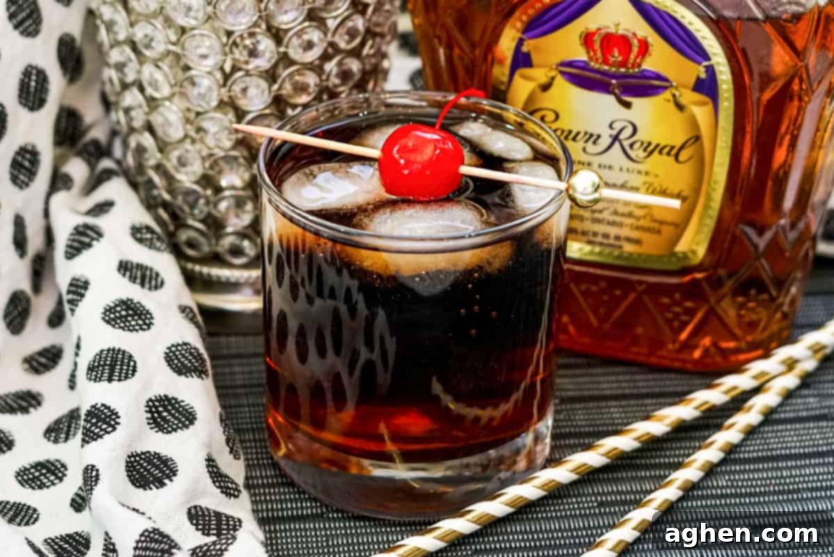 Whiskey Cherry Coke Cocktail in glass with ice cubes and maraschino cherry on skewer, with crown royal bottle behind the glass