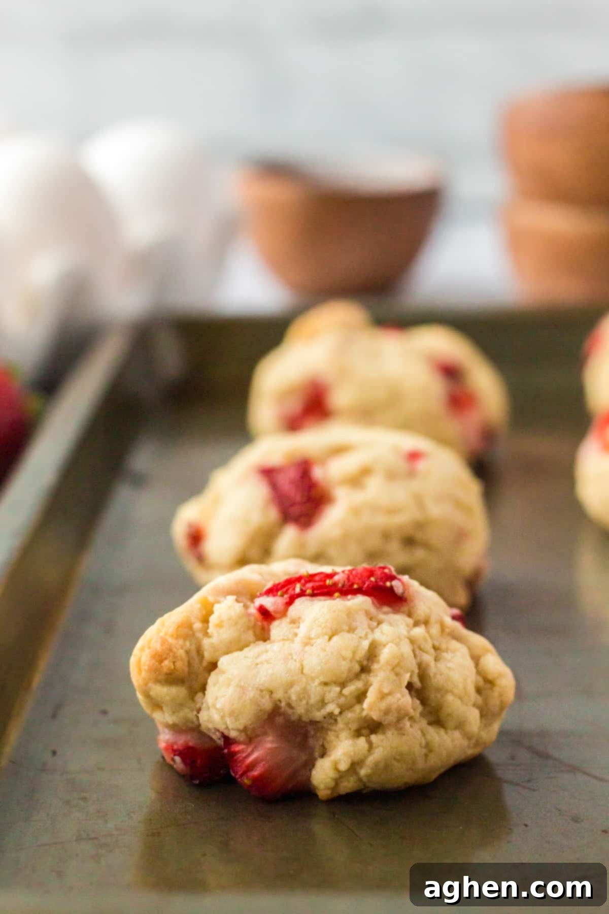 A close-up shot of thick, golden-brown strawberry shortcake cookies fresh from the oven, still on the baking sheet, showcasing their soft, textured tops dotted with strawberry pieces.
