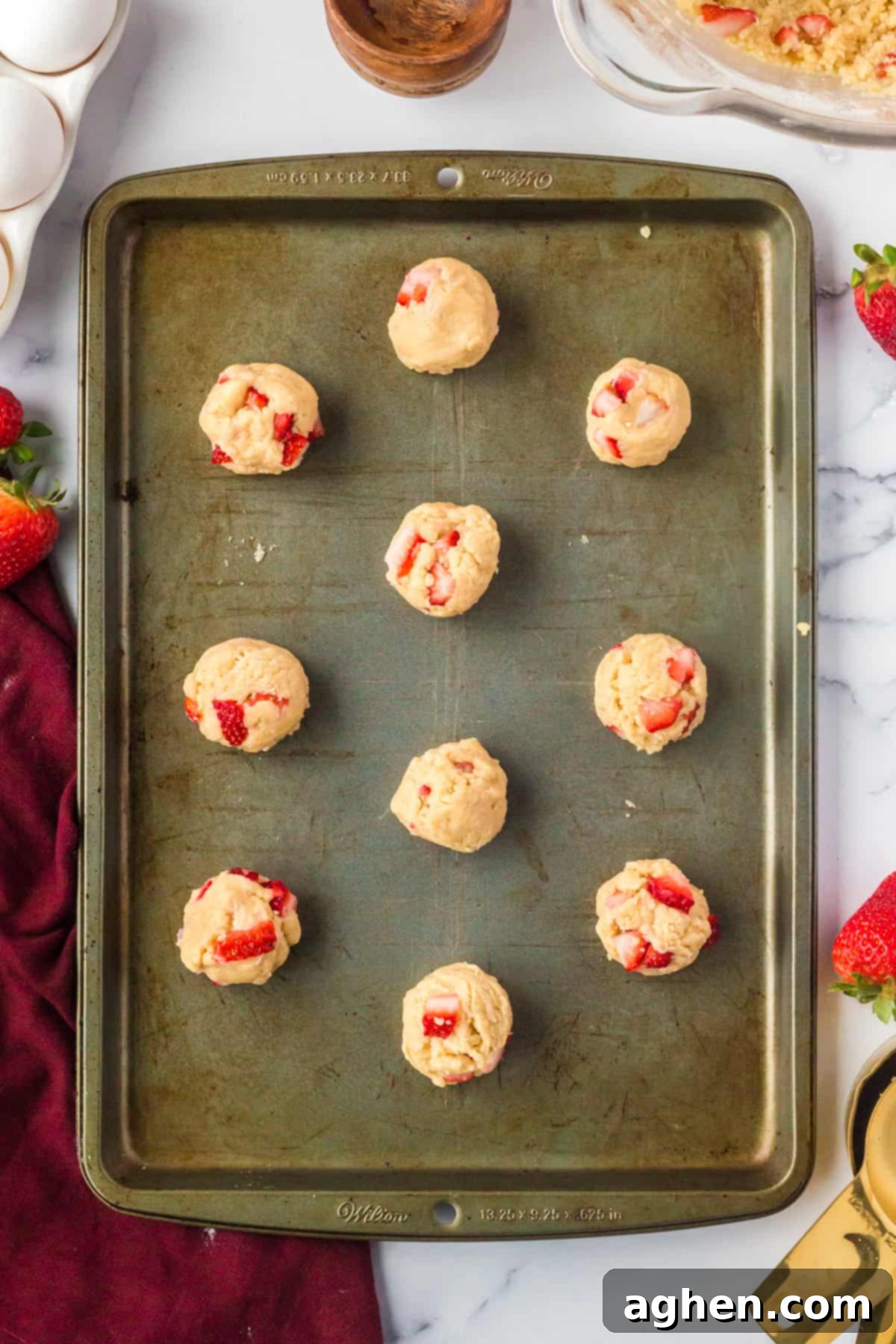 Unbaked strawberry shortcake cookie dough balls, dotted with fresh strawberry pieces, perfectly arranged on a baking sheet lined with parchment paper, ready for the oven.