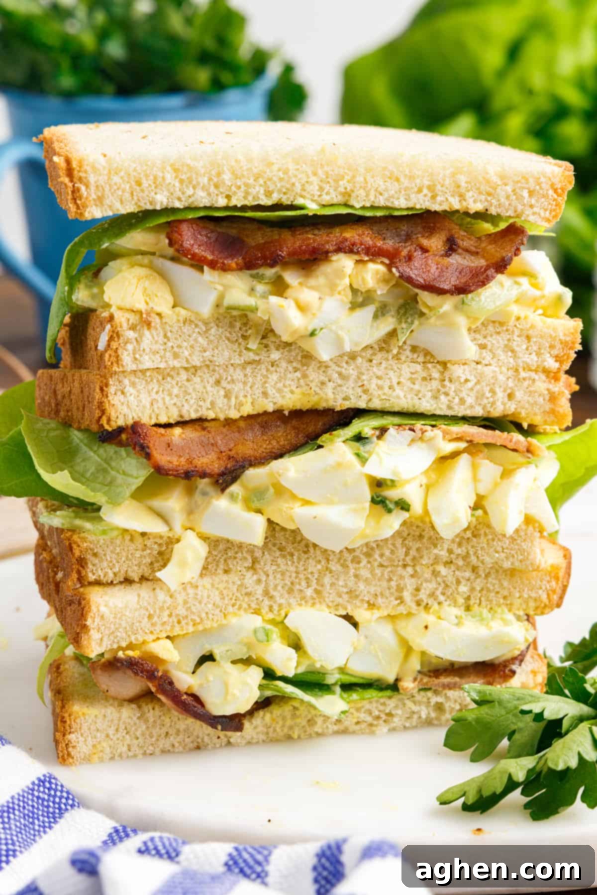 Thee halves of egg salad sandwiches with bacon and lettuce stacked on top of one another on a white plate.