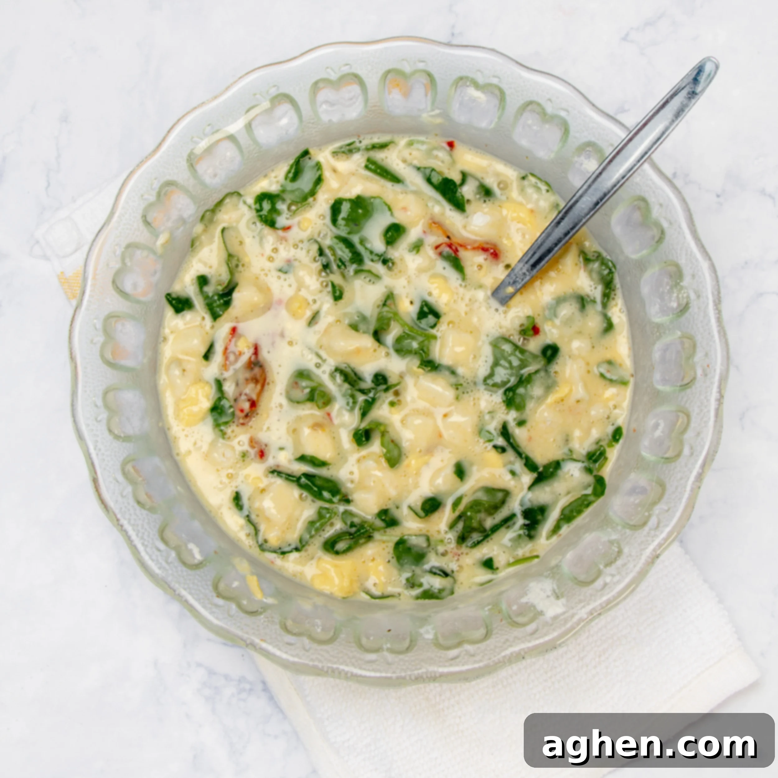 The smooth, whisked egg and milk mixture is being carefully poured into a large mixing bowl filled with the feta, spinach, sun-dried tomatoes, and flour mixture, captured mid-pour just before the final gentle combination to create the muffin batter.