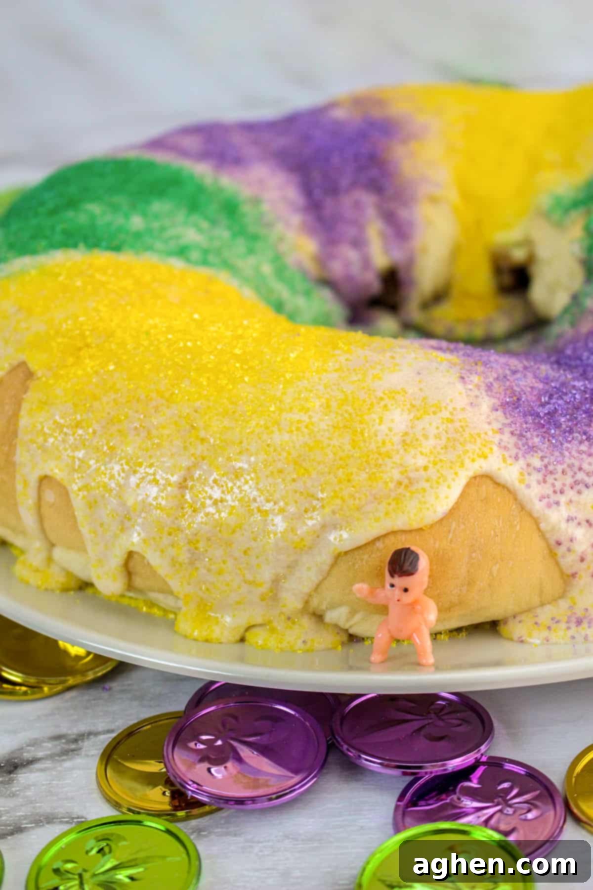Easy homemade king cake with vanilla glaze and decorated with purple, green, and yellow sanding sugar. A small plastic baby in one the white platter next to the cake.