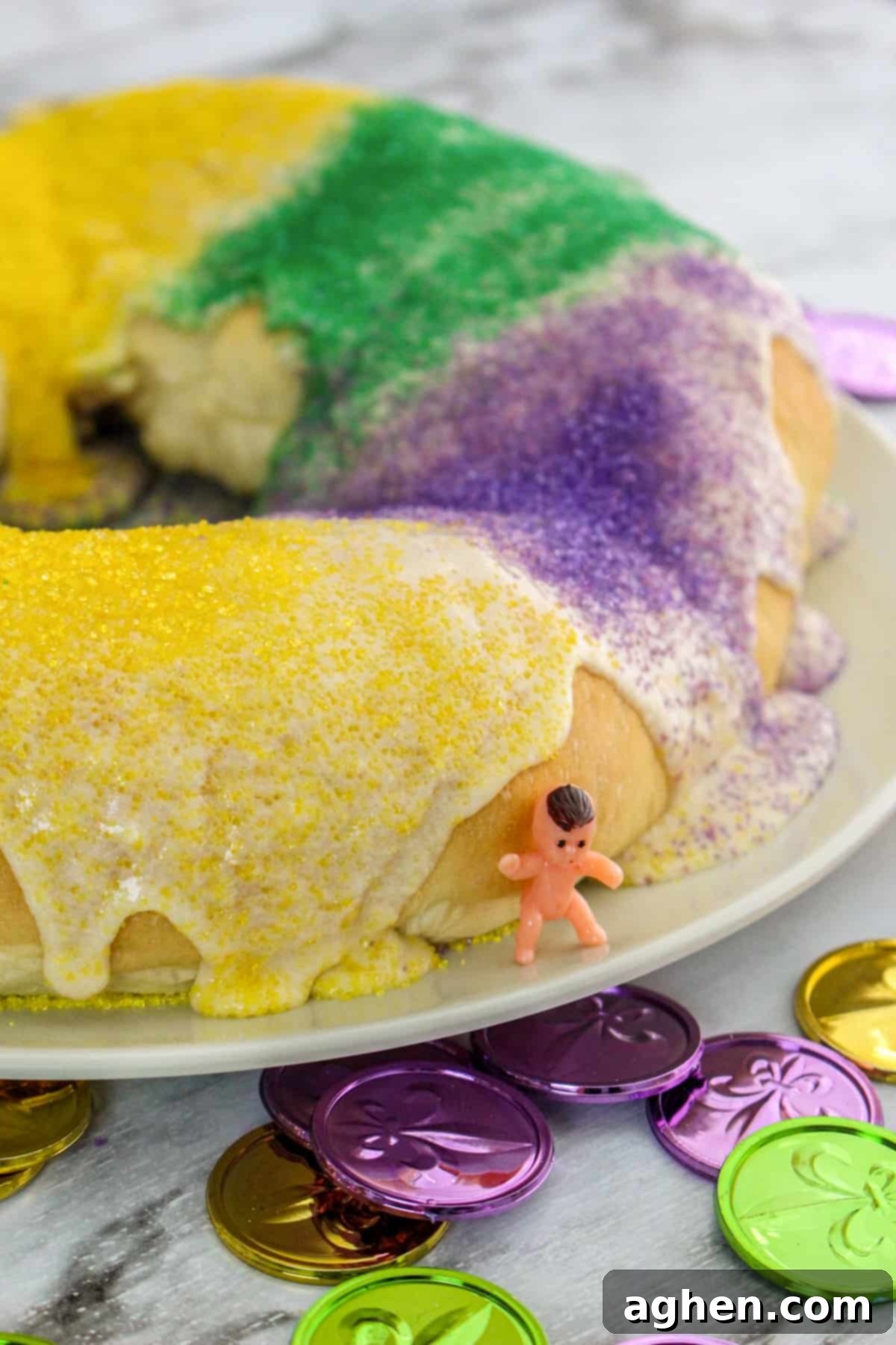 King cake with glaze and green, yellow, and purple sanding sugars. A small toy baby is leaning on the cake.