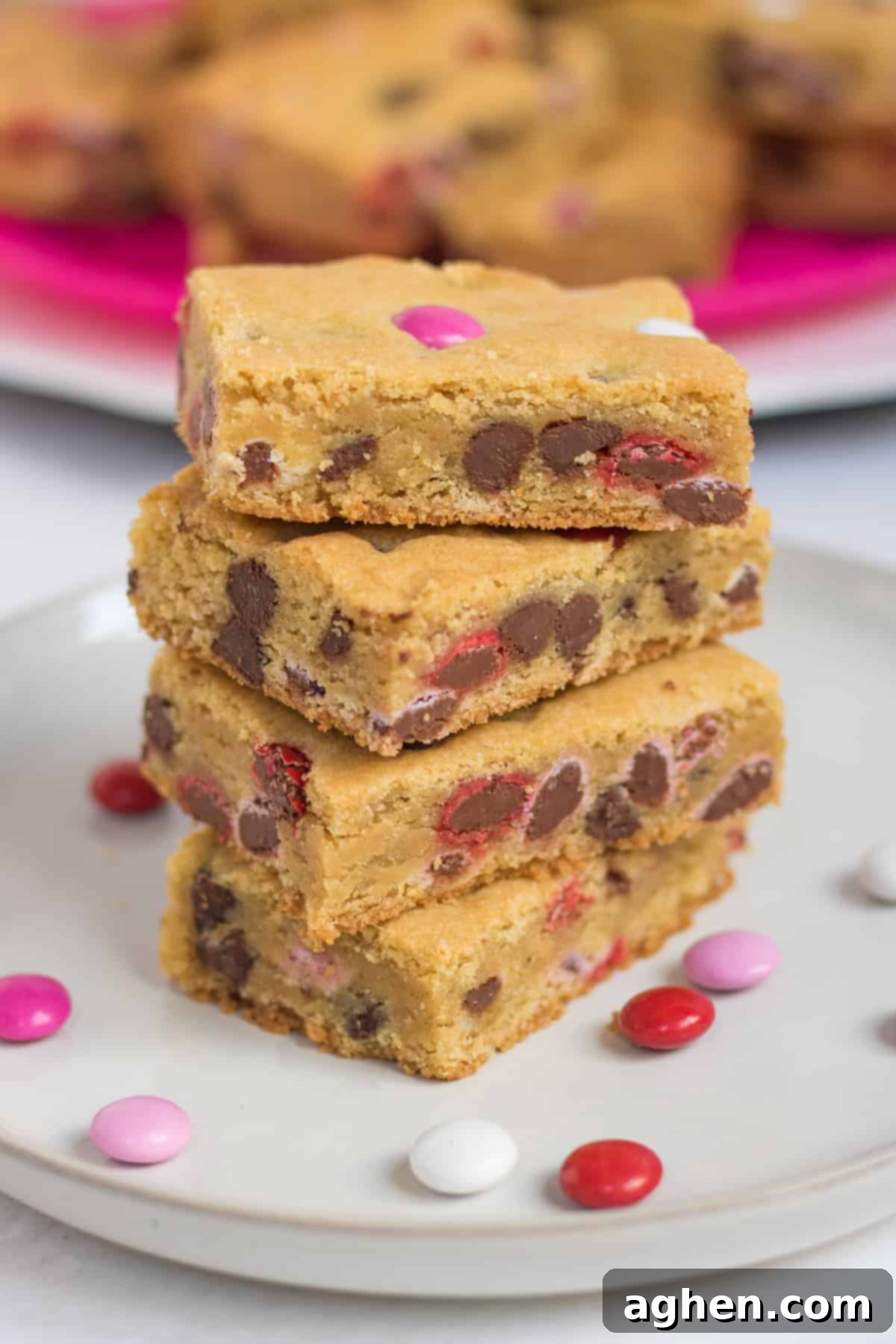 Sweetheart Cookie Squares 2 Four Valentine cookie bars with red, white, and pink M&Ms stacked on top of one another on a white plate, with additional M&Ms sprinkled around them. More cookie bars are visible on a bright pink plate in the background.