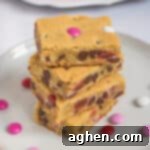 Sweetheart Cookie Squares 11 Four Valentine cookie bars with M&Ms stacked on top of one another on white plate with additional M&Ms sprinkled around them and more bars on bright pink plate in background.