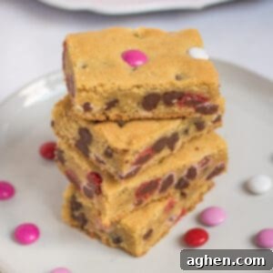 Sweetheart Cookie Squares 10 Four Valentine cookie bars with M&Ms stacked on top of one another on white plate with additional M&Ms sprinkled around them and more bars on bright pink plate in background.