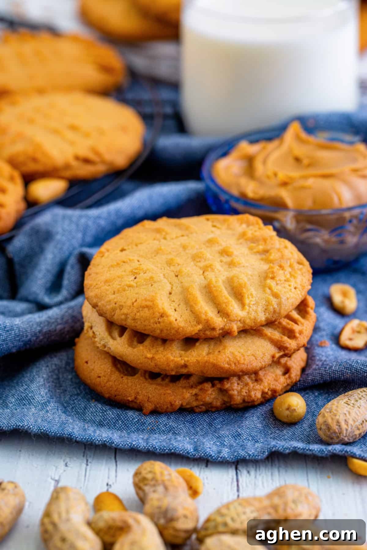 A delightful stack of three 'Lunch Lady' old-fashioned peanut butter cookies, surrounded by more cookies, a jar of peanut butter, peanuts, and a refreshing glass of milk.