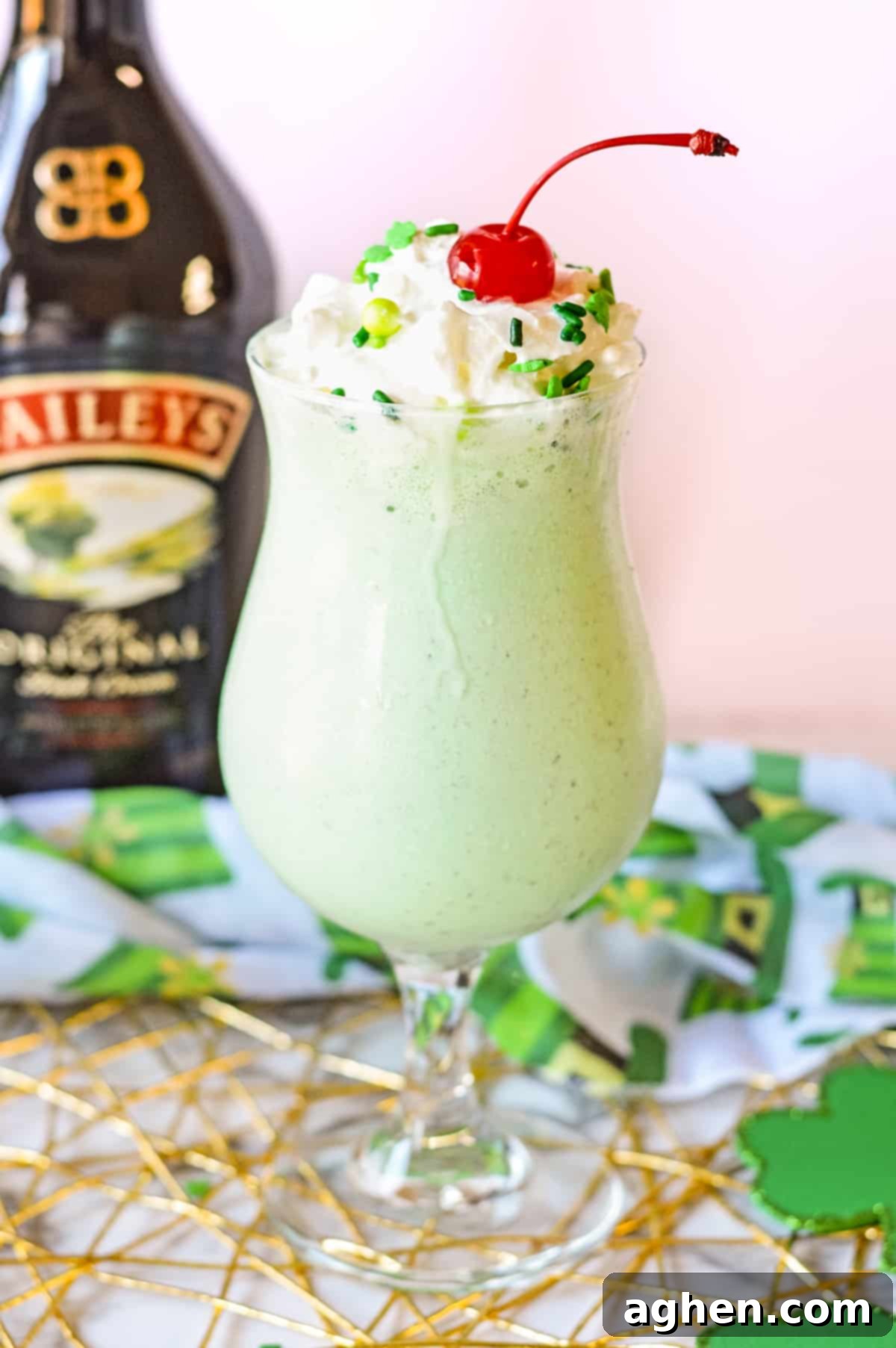 Vibrant mint green Boozy Shamrock Shake with fluffy whipped cream, green sprinkles, and a cherry. A bottle of Baileys in the soft-focus background highlights the adult nature of the drink.
