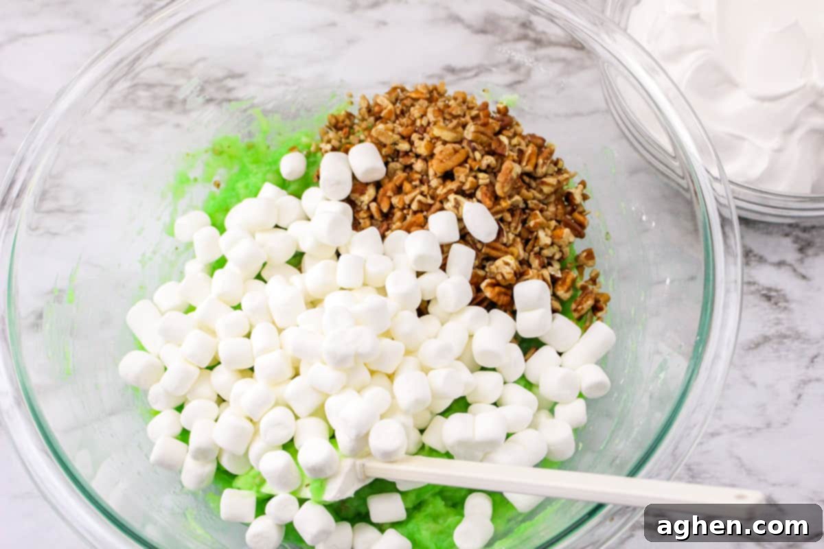 Mini marshmallows and chopped pecans added on top of the pistachio pudding mixture in a mixing bowl, ready to be stirred.