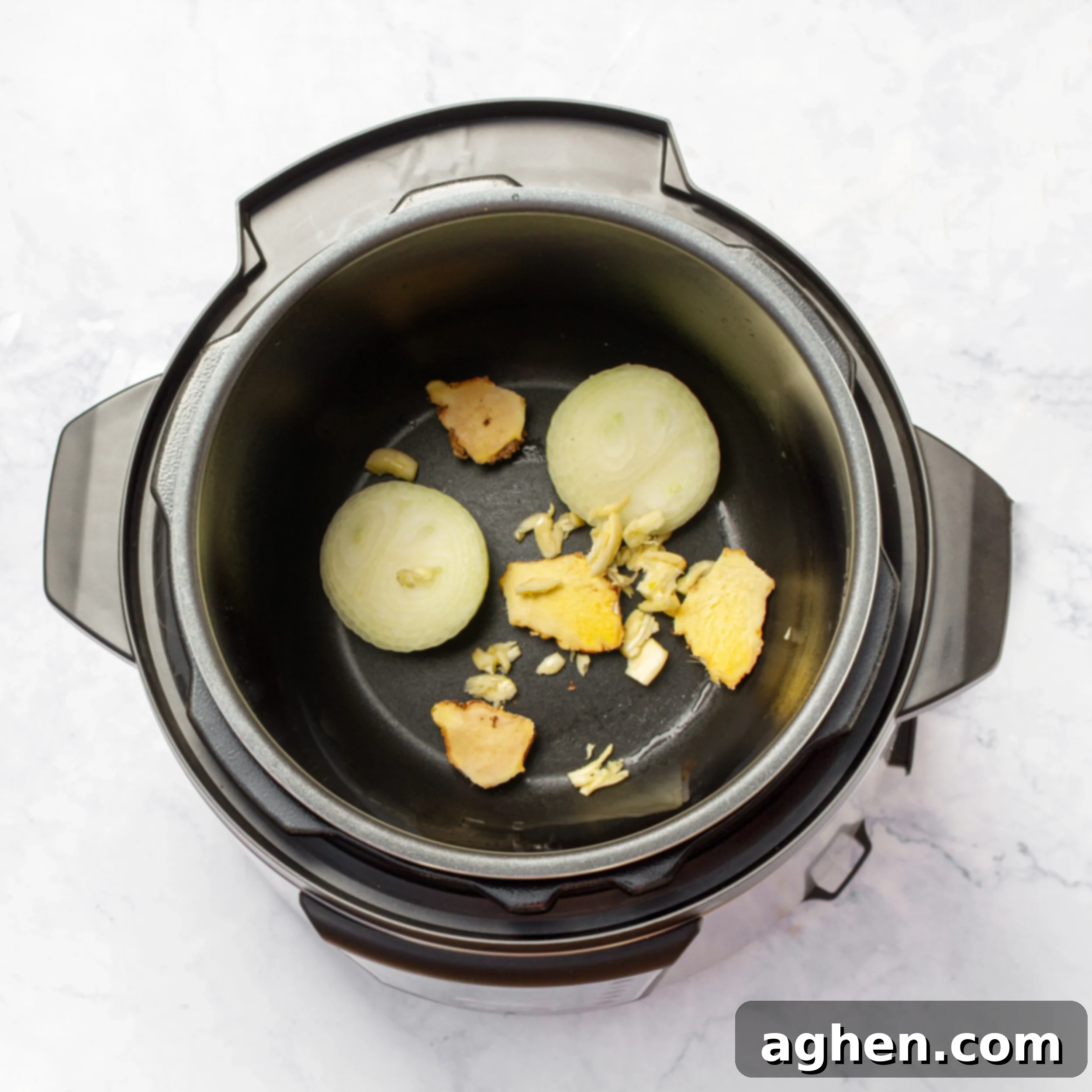 Halved onion, crushed garlic, and whole ginger root at the bottom of an Instant Pot.