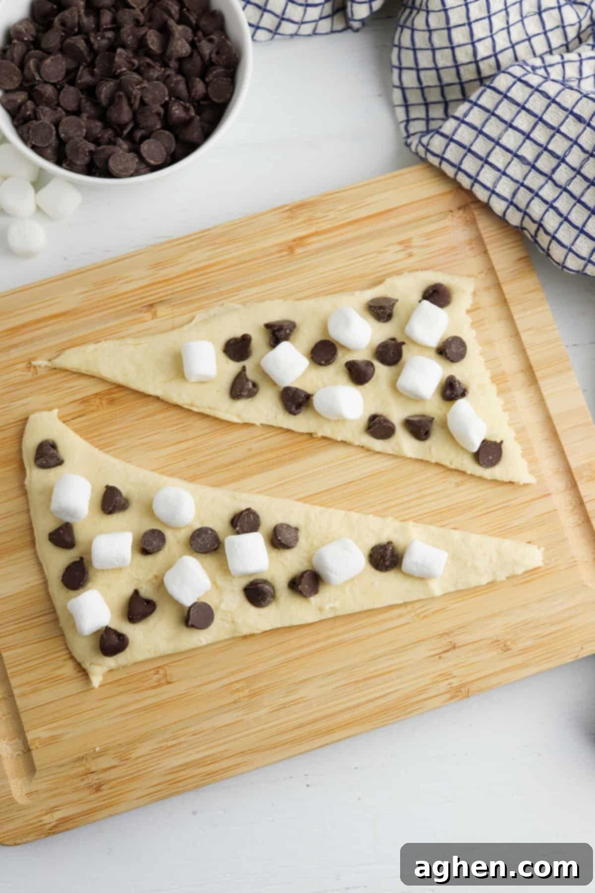 Two triangles of crescent roll dough laid flat on a cutting board and topped with mini marshmallows and chocolate chips.