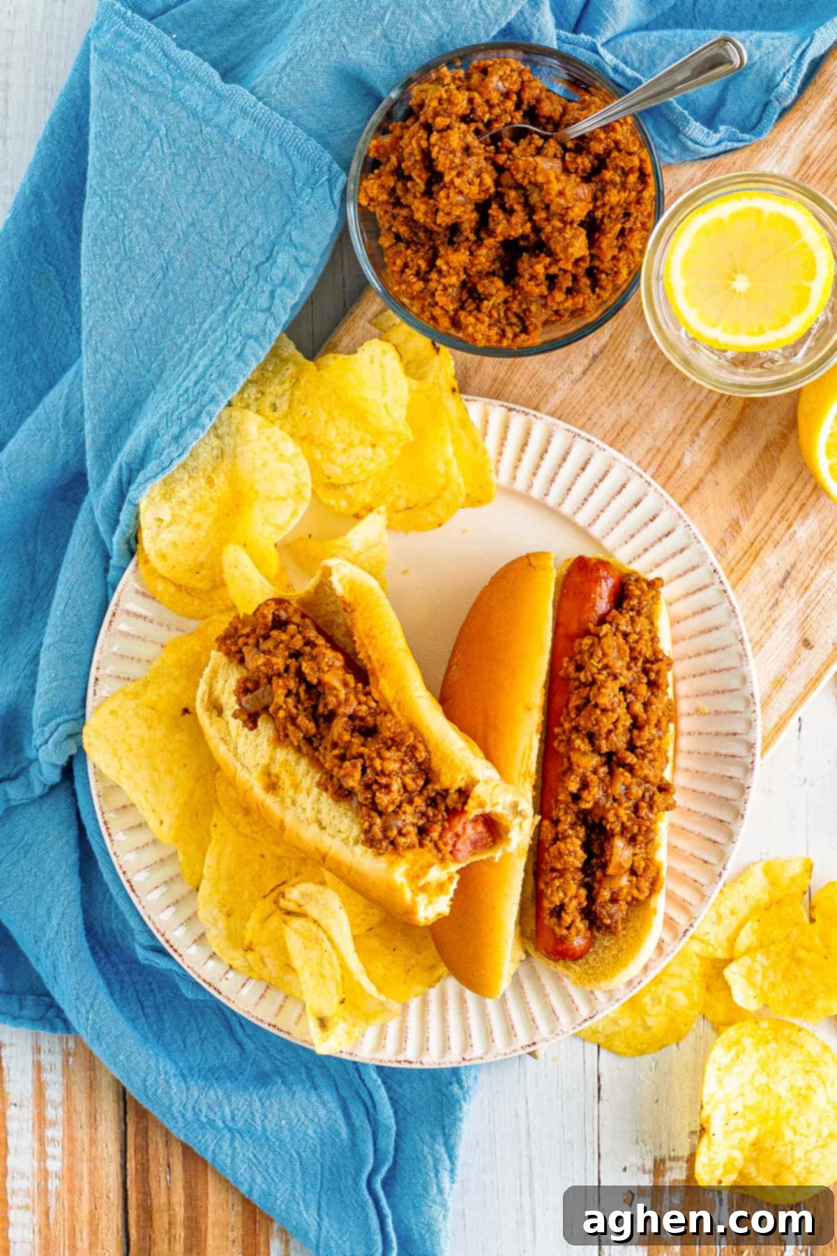 Hot dogs with chili sauce served with potato chips. More chili can be seen in background.
