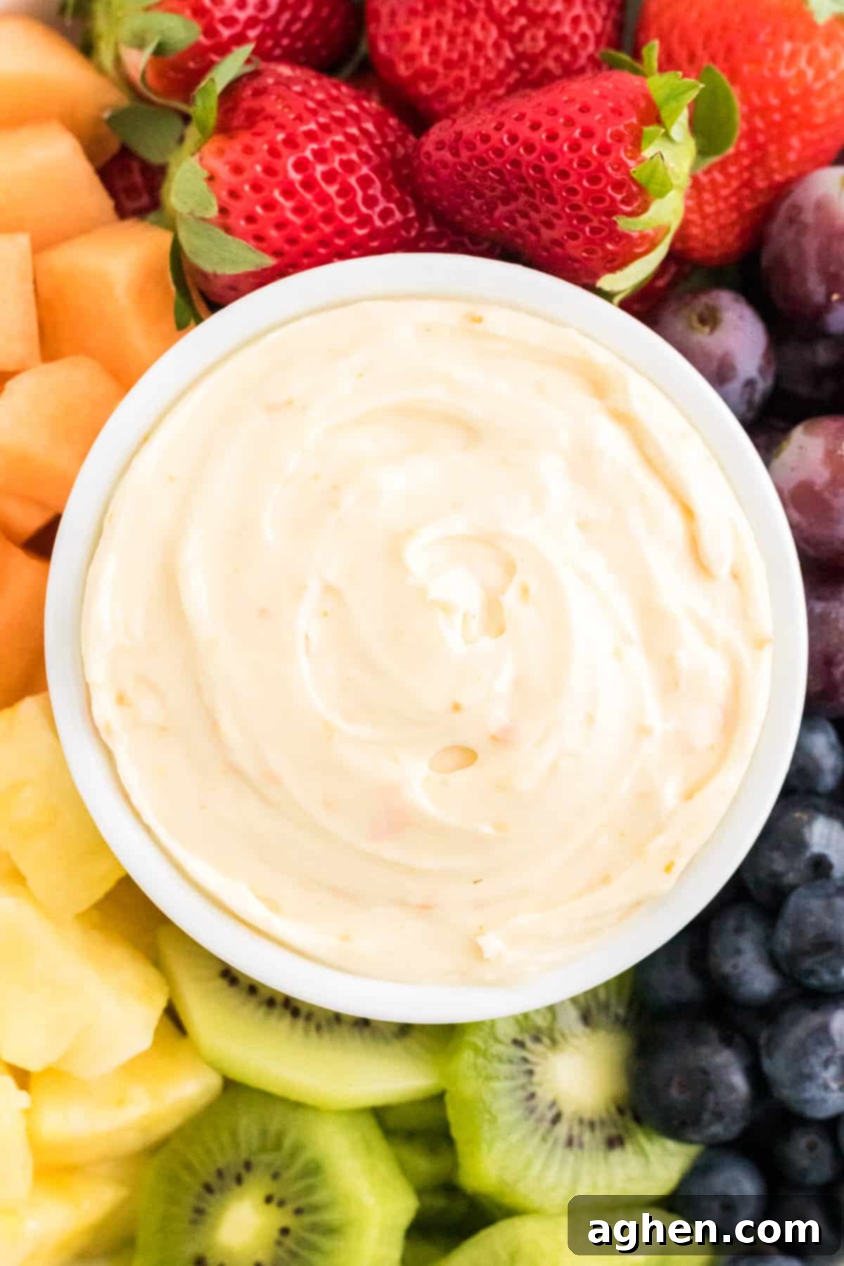 Cream cheese fruit dip in white bowl served with fresh strawberries, pineapple, grapes, kiwi, blueberries, and cantaloupe.