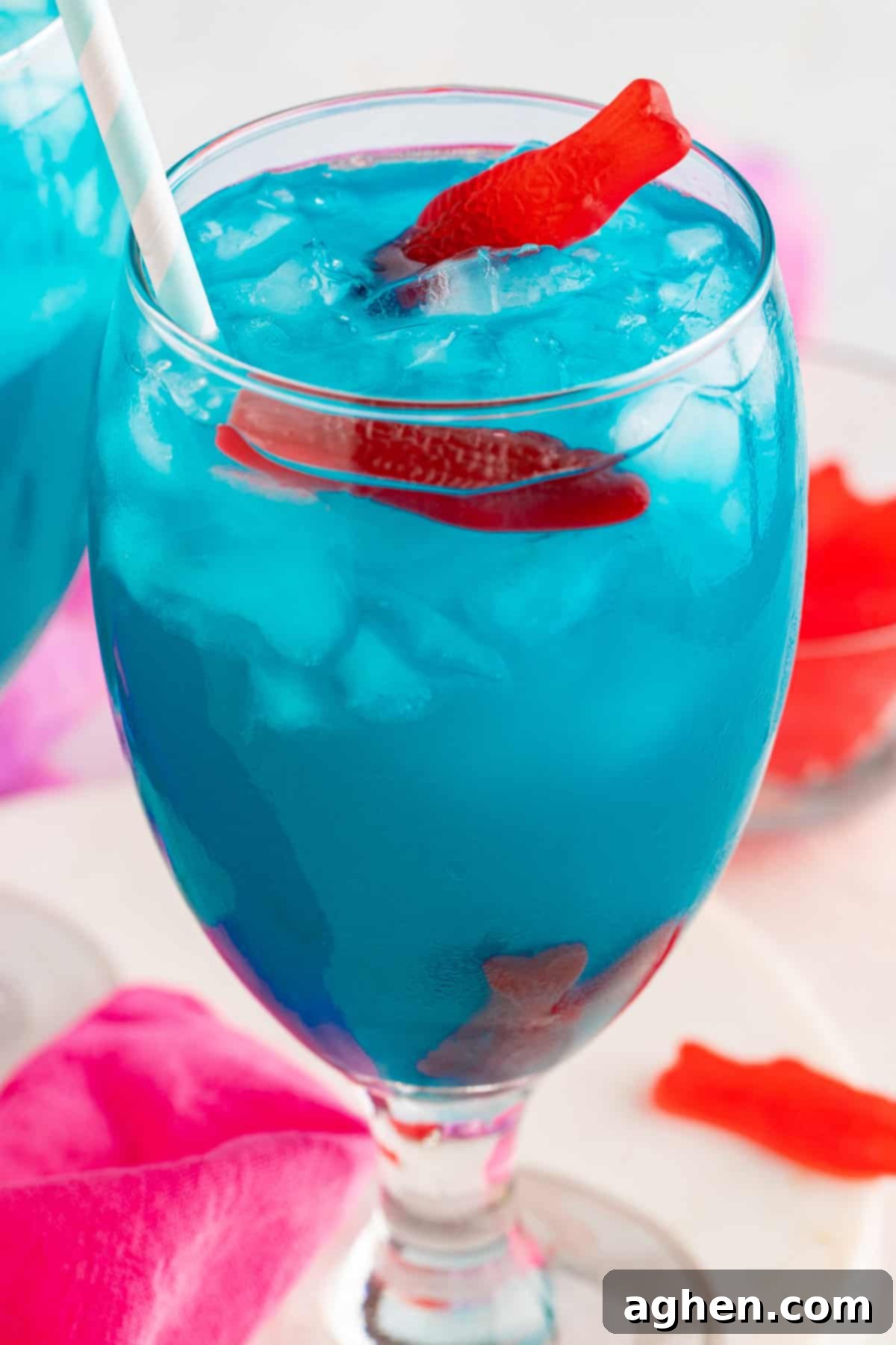 Blue ocean water cocktail in a wine glass with red candy fish sticking out of the top, garnished with a pineapple wedge.