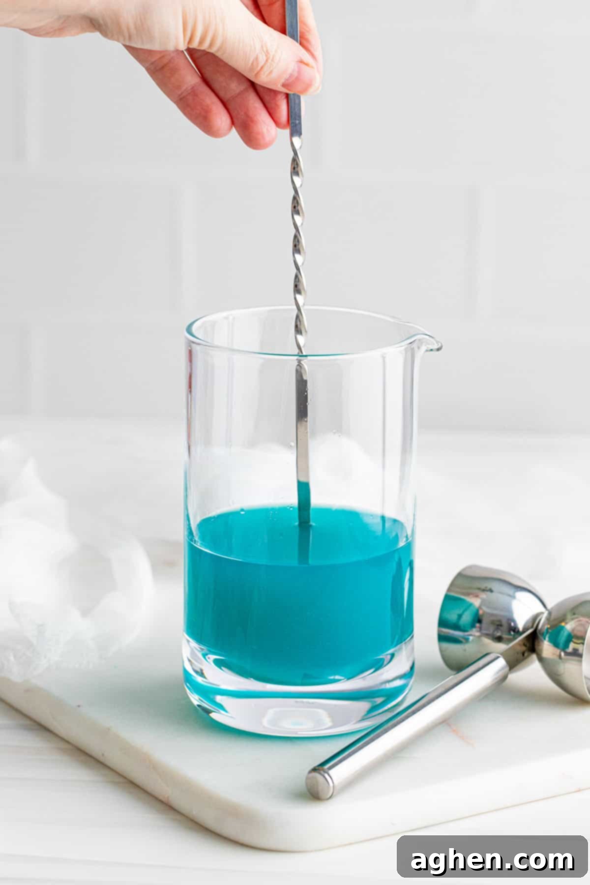 A blue cocktail being stirred gently in a large mixing glass with a bar spoon.