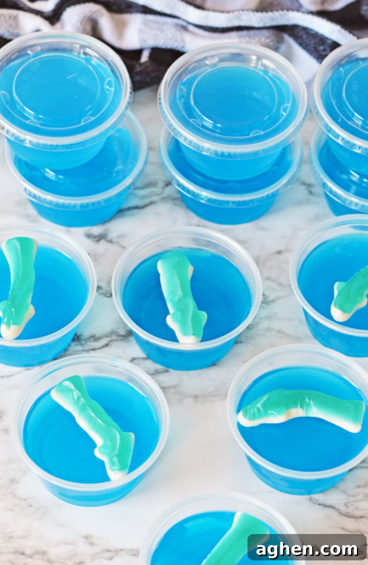 Collection of bright blue gummy shark jello shots arranged in clear 2-ounce plastic shot glasses, some with their corresponding lids.