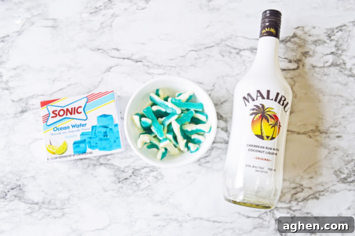A flat lay of the key ingredients for shark jello shots: a box of Sonic Ocean Water jello, a bowl of blue shark gummy candies, and a bottle of Malibu coconut rum.