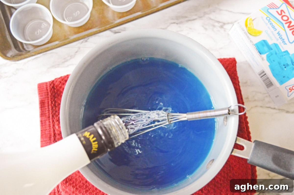 A bottle of Malibu coconut rum being poured into the bright blue jello mixture, preparing the base for shark shots.