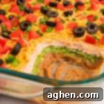 Seven Layer Taco Fiesta 11 Partially scooped 7 layer dip without cream cheese, highlighting colorful layers of bean dip, sour cream, and guacamole, and garnished with fresh tomatoes, olives, and green onions