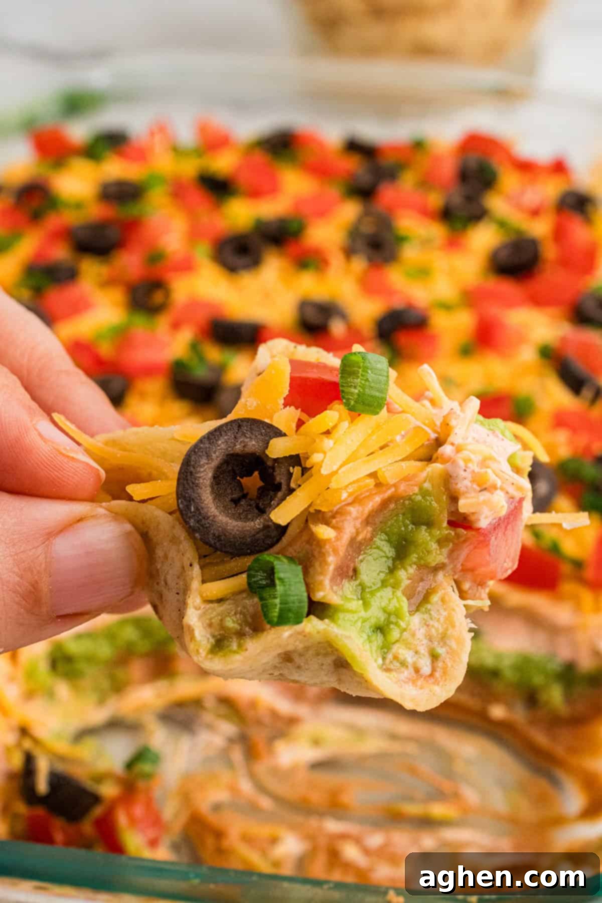 Seven Layer Taco Fiesta 3 Close-up of a tortilla chip loaded with 7-layer taco dip without cream cheese, featuring layers of refried beans, guacamole, sour cream, shredded cheese, tomatoes, black olives, and green onions.
