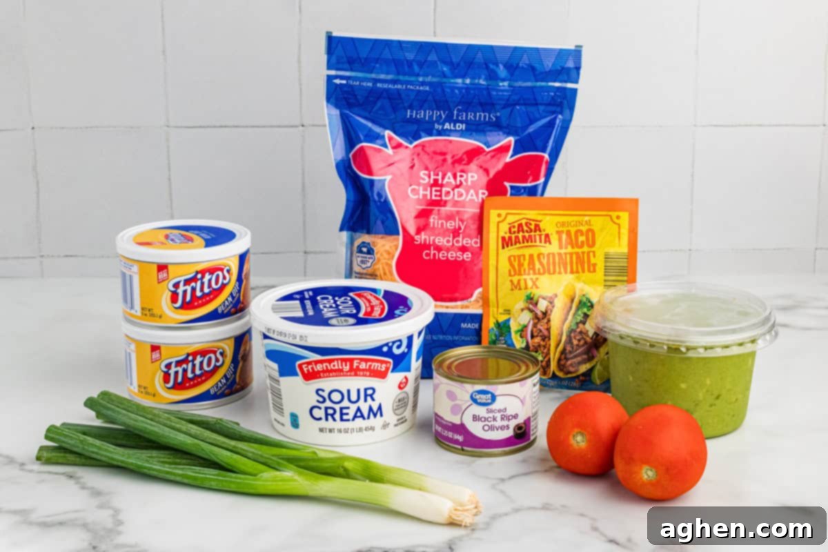 Seven Layer Taco Fiesta 4 Ingredients for a 7-layer taco dip arranged on a countertop, including Fritos bean dip, sour cream, shredded sharp cheddar cheese, taco seasoning mix, guacamole, sliced black olives, tomatoes, and green onions.