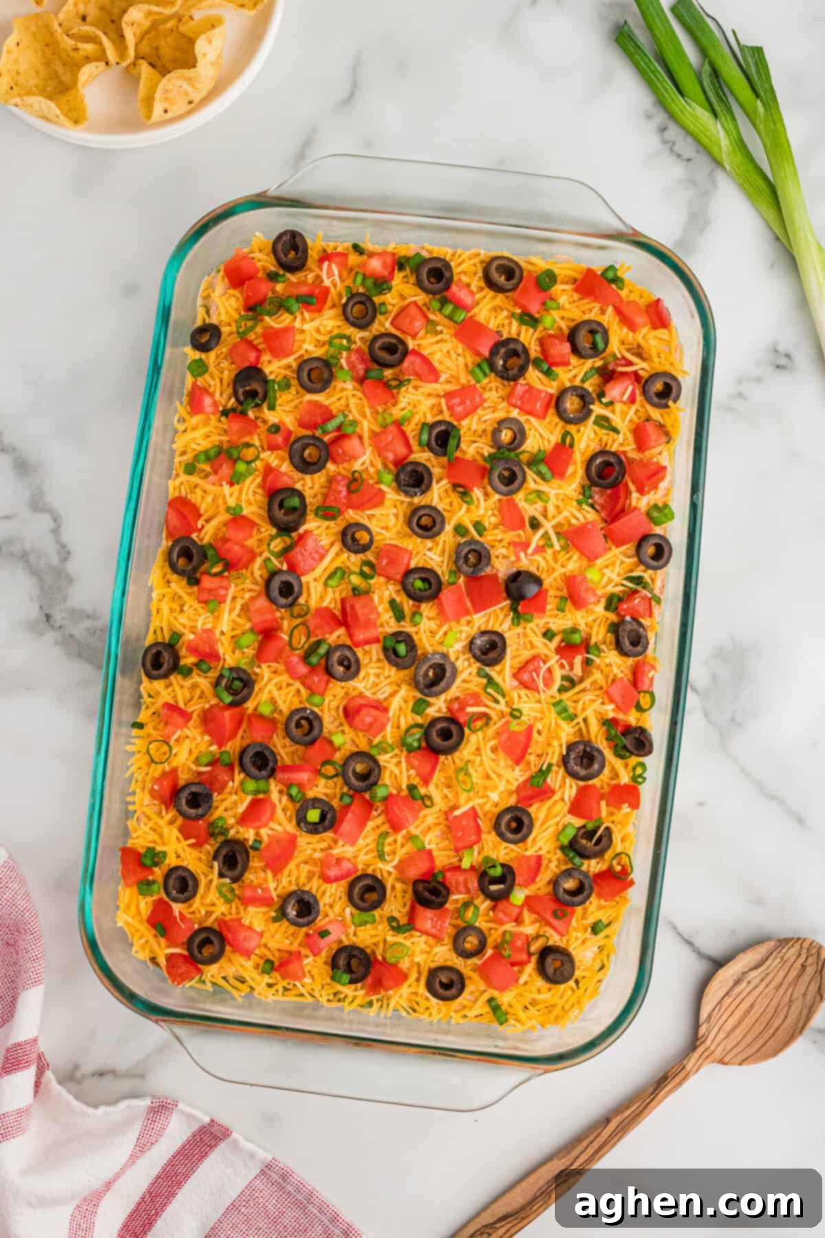 Seven Layer Taco Fiesta 6 9 x 13 glass dish filled with a 7-layer taco dip made without cream cheese, topped with shredded cheddar, diced tomatoes, black olives, and green onions.