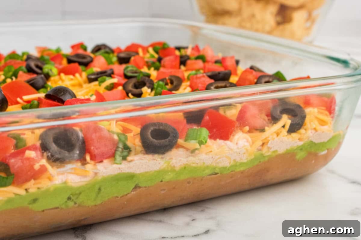 Seven Layer Taco Fiesta 7 Side view of a 9x13 dish of 7-layer dip, showing distinct layers of refried beans, guacamole, taco-seasoned sour cream, shredded cheese, and toppings like tomatoes, olives, and green onions.