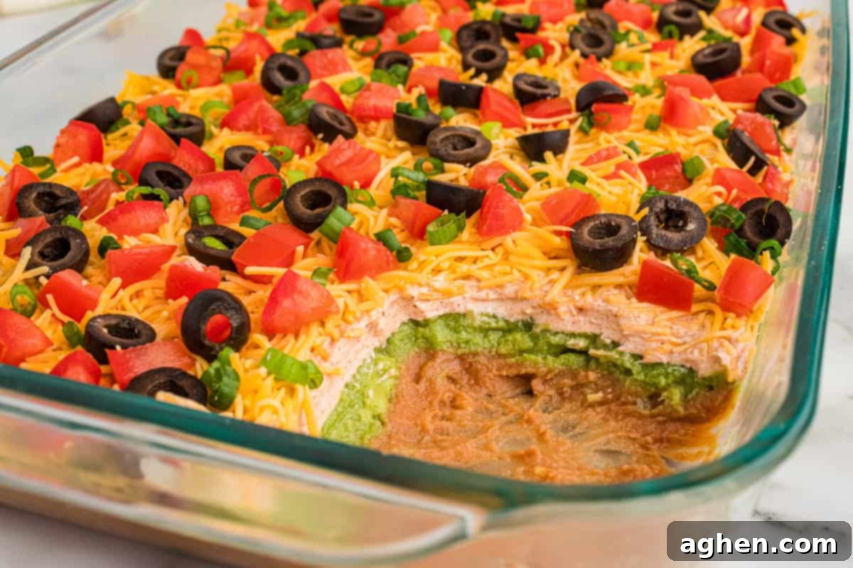 Seven Layer Taco Fiesta 9 Close-up of a partially scooped 7-layer dip in a glass dish, showcasing layers of refried beans, guacamole, seasoned sour cream, shredded cheddar cheese, and toppings of diced tomatoes, black olives, and green onions.