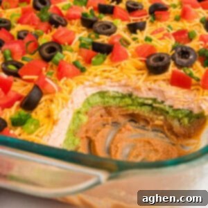 Seven Layer Taco Fiesta 10 Partially scooped 7 layer dip without cream cheese, highlighting colorful layers of bean dip, sour cream, and guacamole, and garnished with fresh tomatoes, olives, and green onions