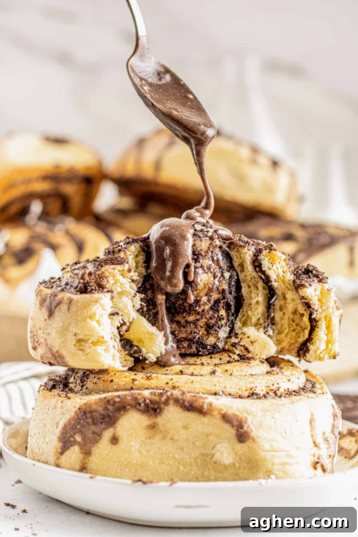 Cookies and Cream Dream Buns 3 Spoon drizzling oreo glaze over large cinnamon rolls in a baking pan, showcasing the rich texture of the frosting.