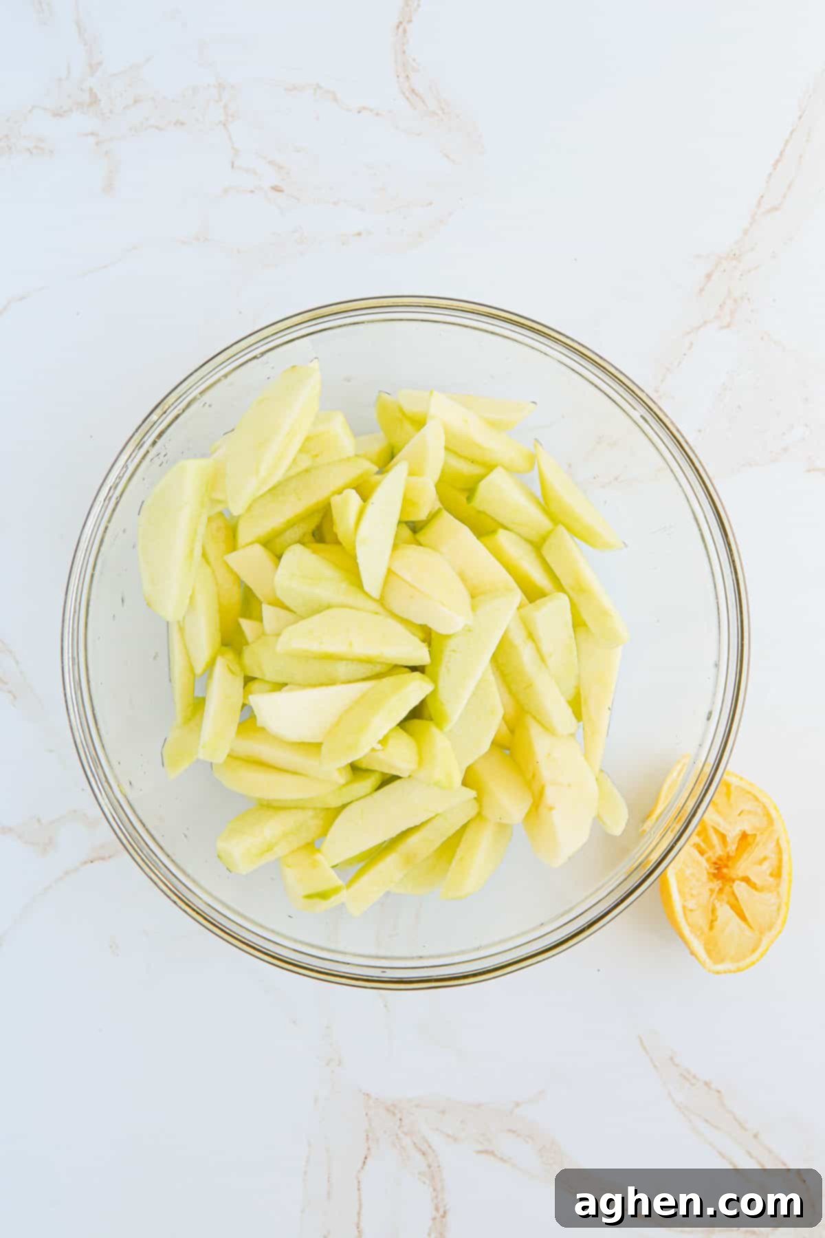 Peeled, cored, and sliced apples in a mixing bowl, glistening with lemon juice to prevent browning. A squeezed lemon half rests beside it.