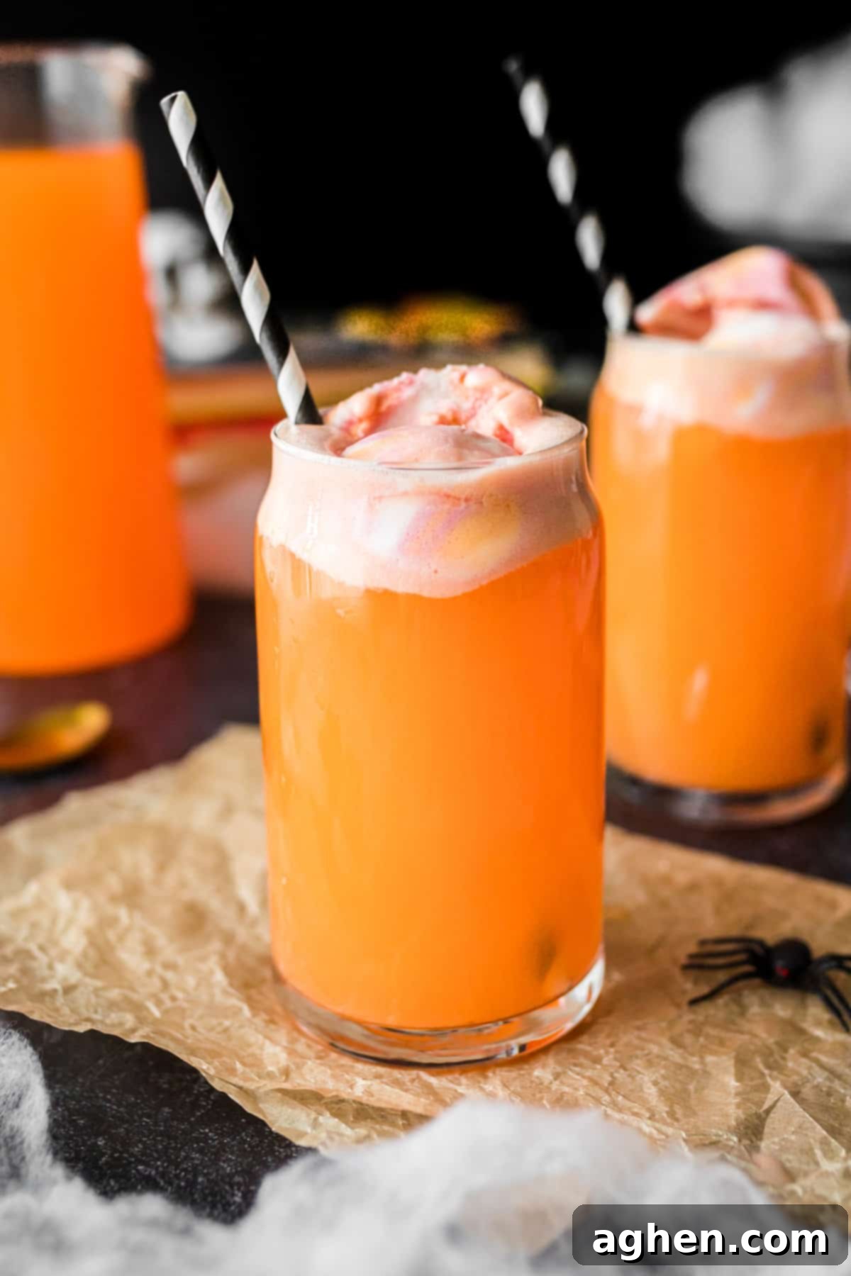 Orange Halloween punch in tall glass topped with sherbet. Another filled glass and pitcher of the punch are in the background.