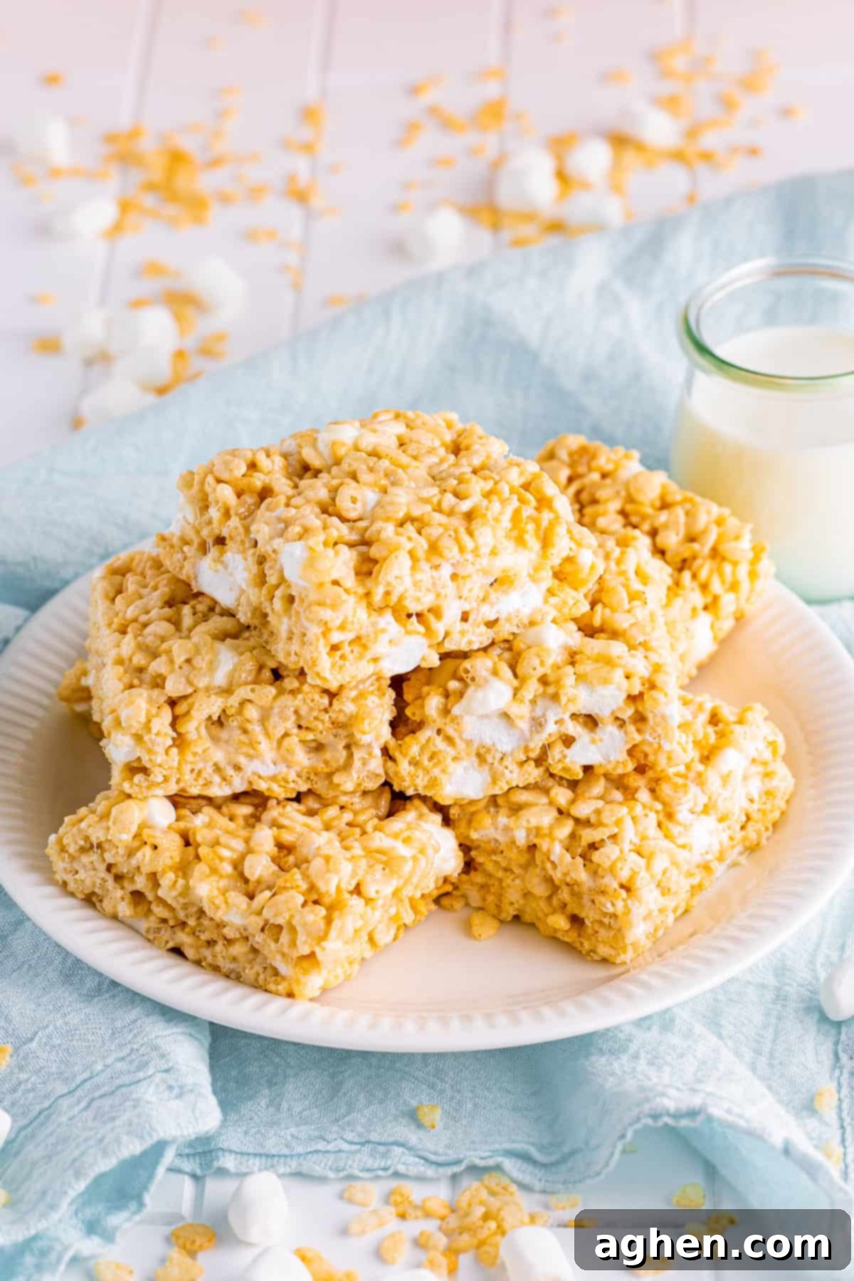Thick, chewy, and extra gooey Rice Krispies Treats stacked on a white plate with a glass of milk in the background, showcasing their irresistible texture.