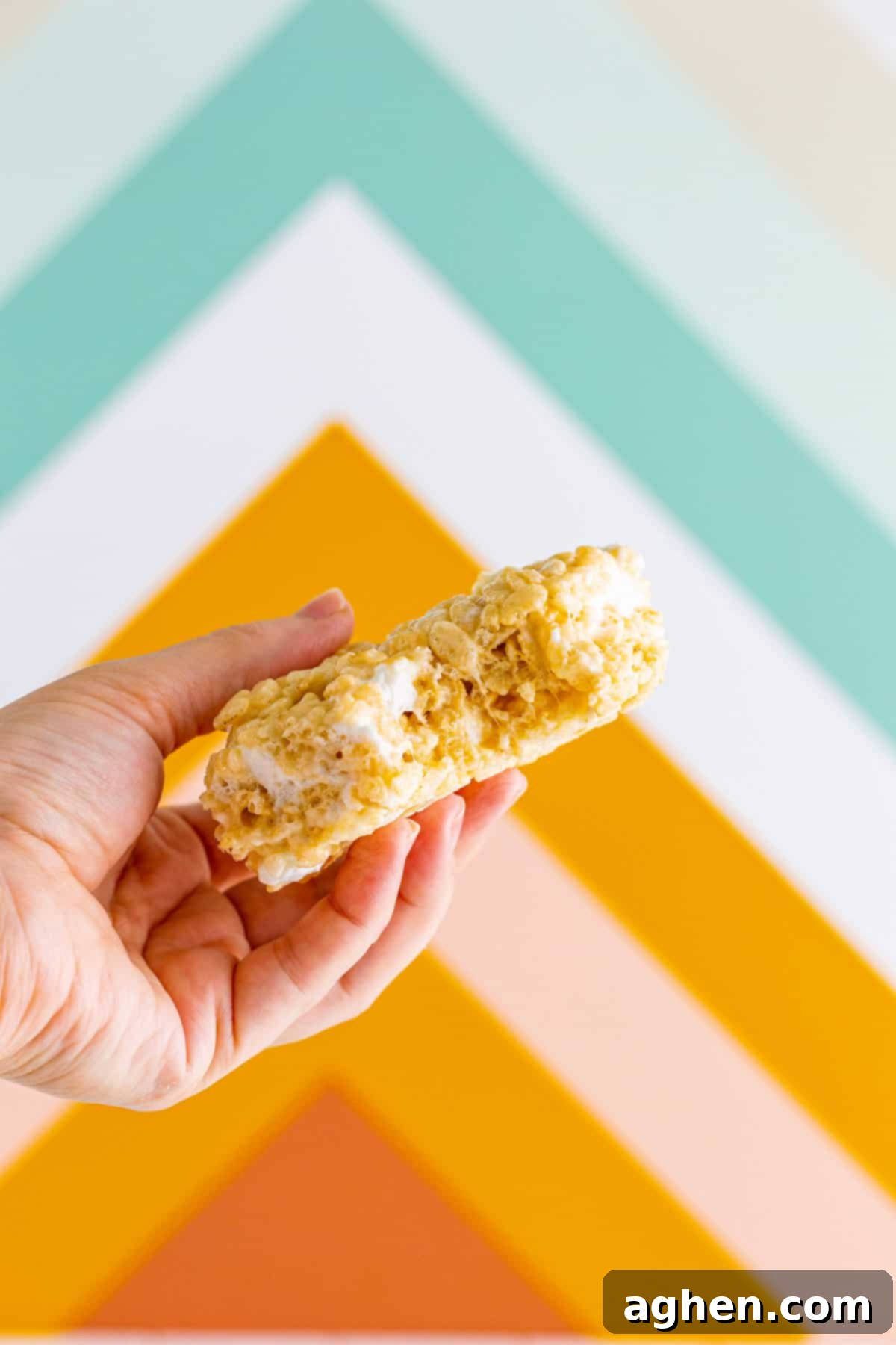 A hand holding a jumbo-sized, bakery-style Rice Krispie treat with a bite taken out, showcasing its incredibly soft and chewy texture.