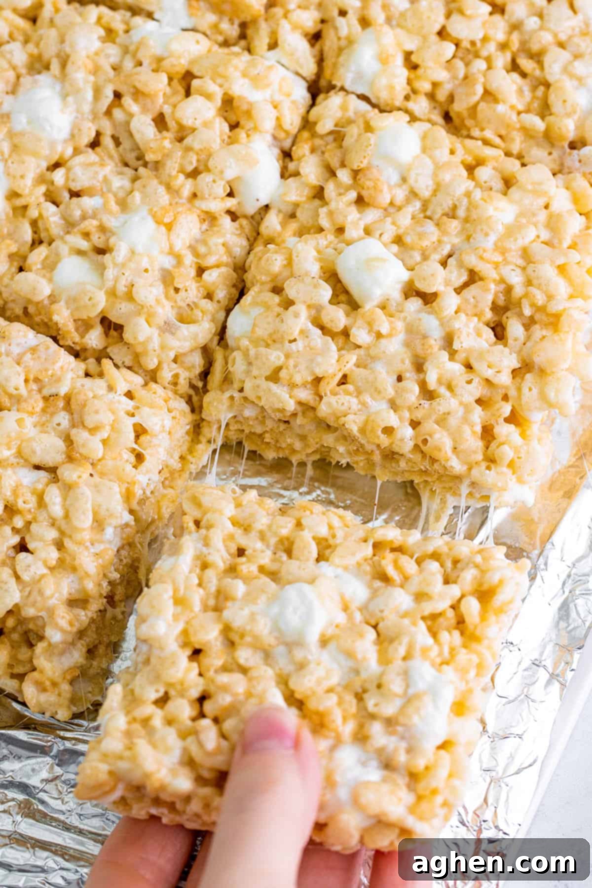 A deliciously gooey Rice Krispie treat being pulled from a tray of freshly made treats, showcasing its irresistible stretch and texture.