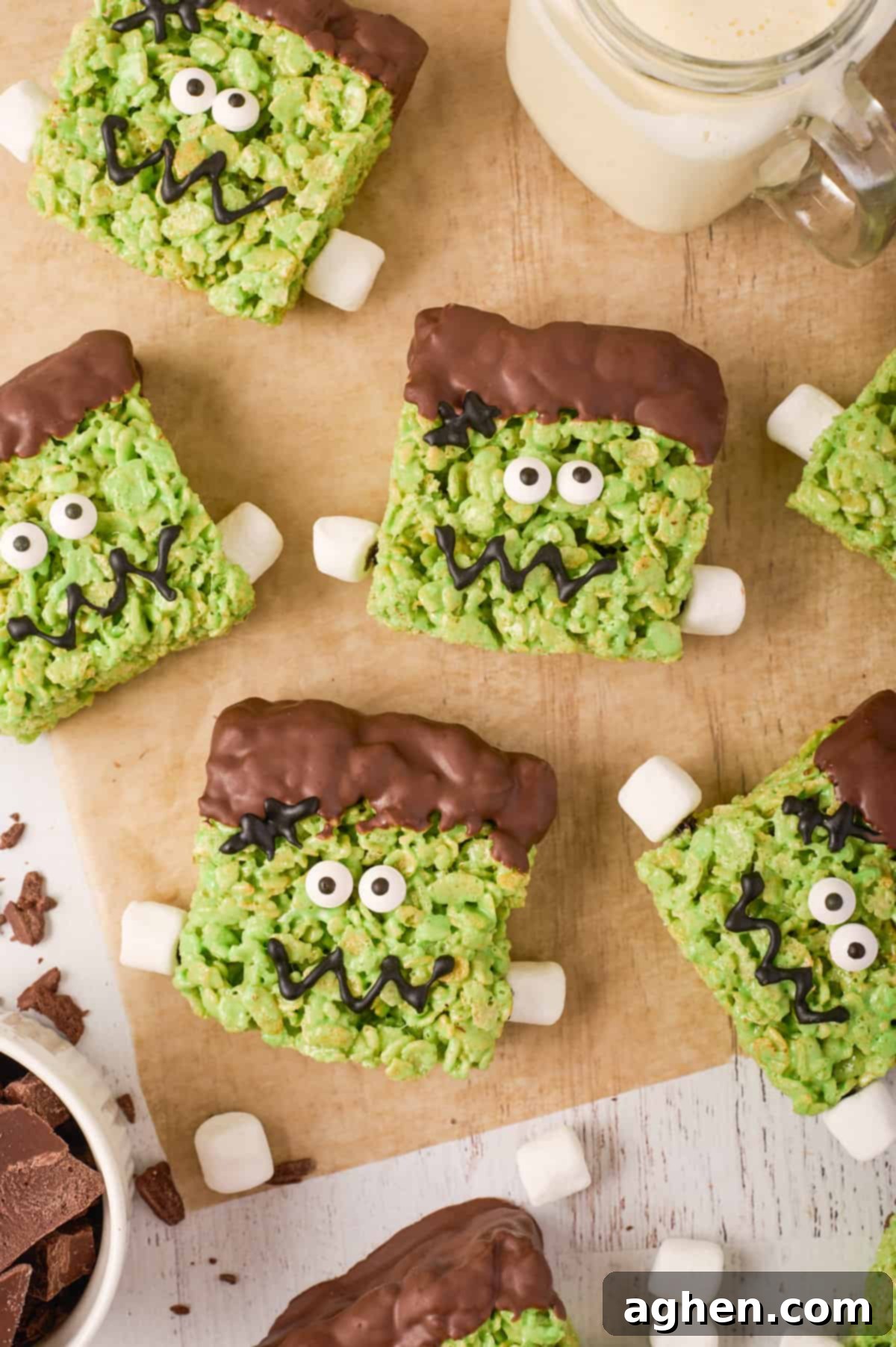 Cute frankenstein rice krispies treats on a piece of parchment paper with mini marshmallows scattered and a glass of milk.