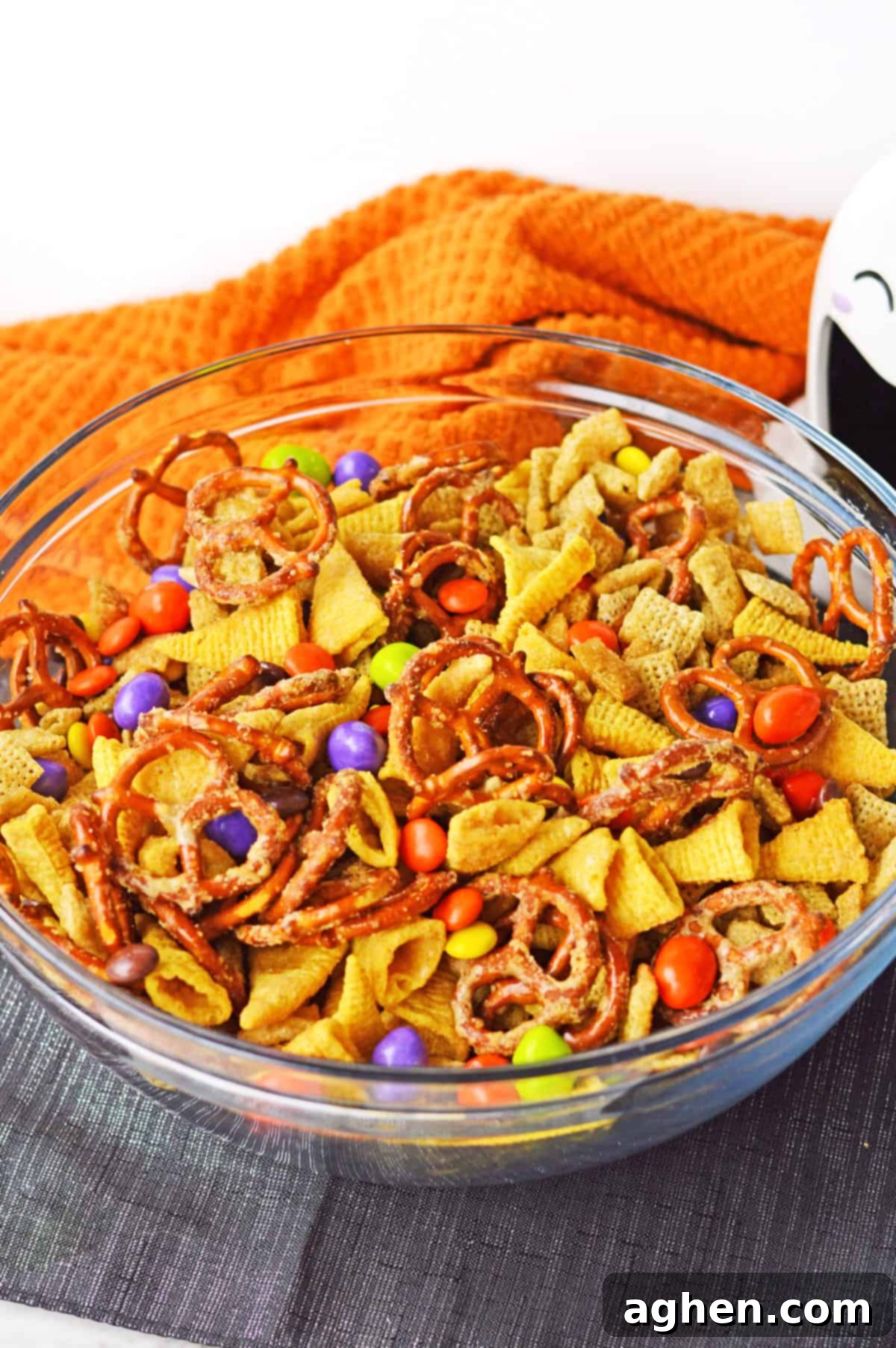 Festive Halloween Chex Mix in a clear glass bowl, surrounded by spooky Halloween decorations like mini pumpkins and spiderwebs.