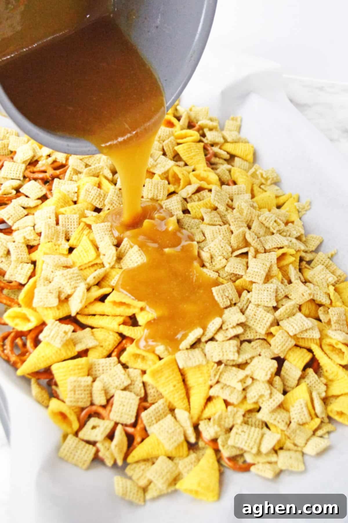 Warm caramel sauce being poured from a saucepan over the Chex mix ingredients on a parchment-lined baking sheet.