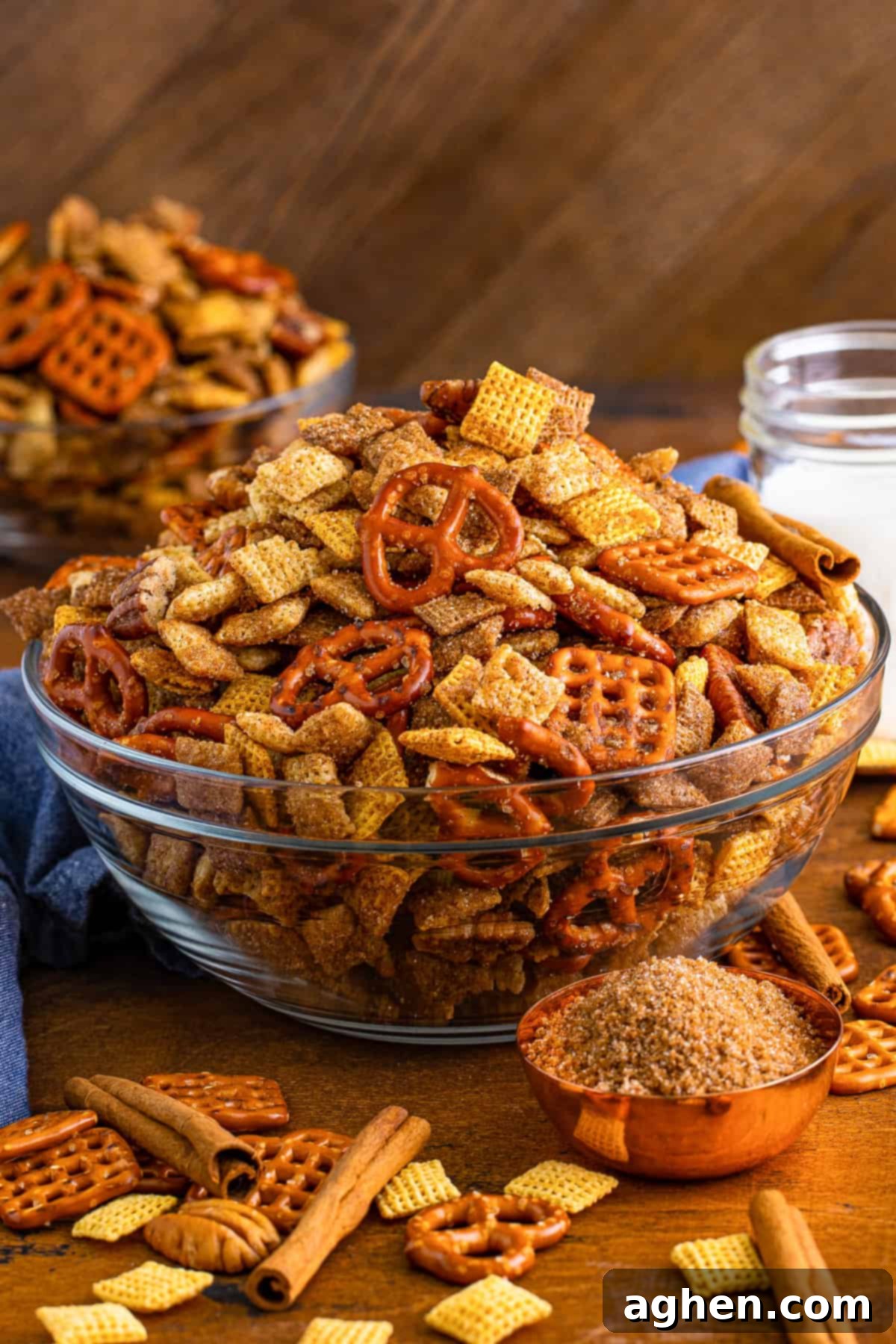 A glass serving bowl overflowing with golden-brown Cinnamon Sugar Chex Mix, with a measuring cup of brown sugar and cinnamon sticks artfully placed beside it.