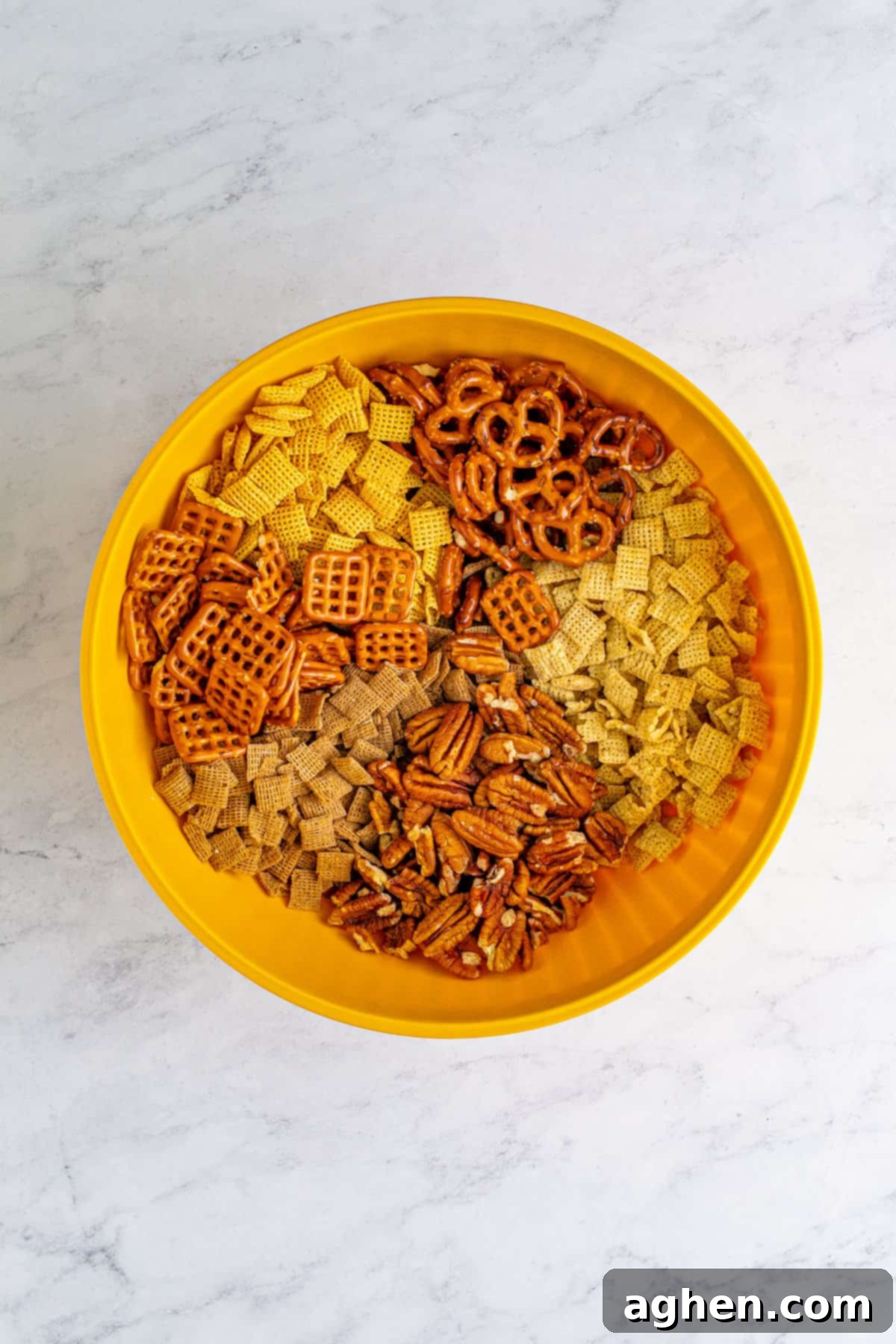A yellow mixing bowl filled with a blend of Chex cereal, pecans, and pretzels, ready for the sauce.